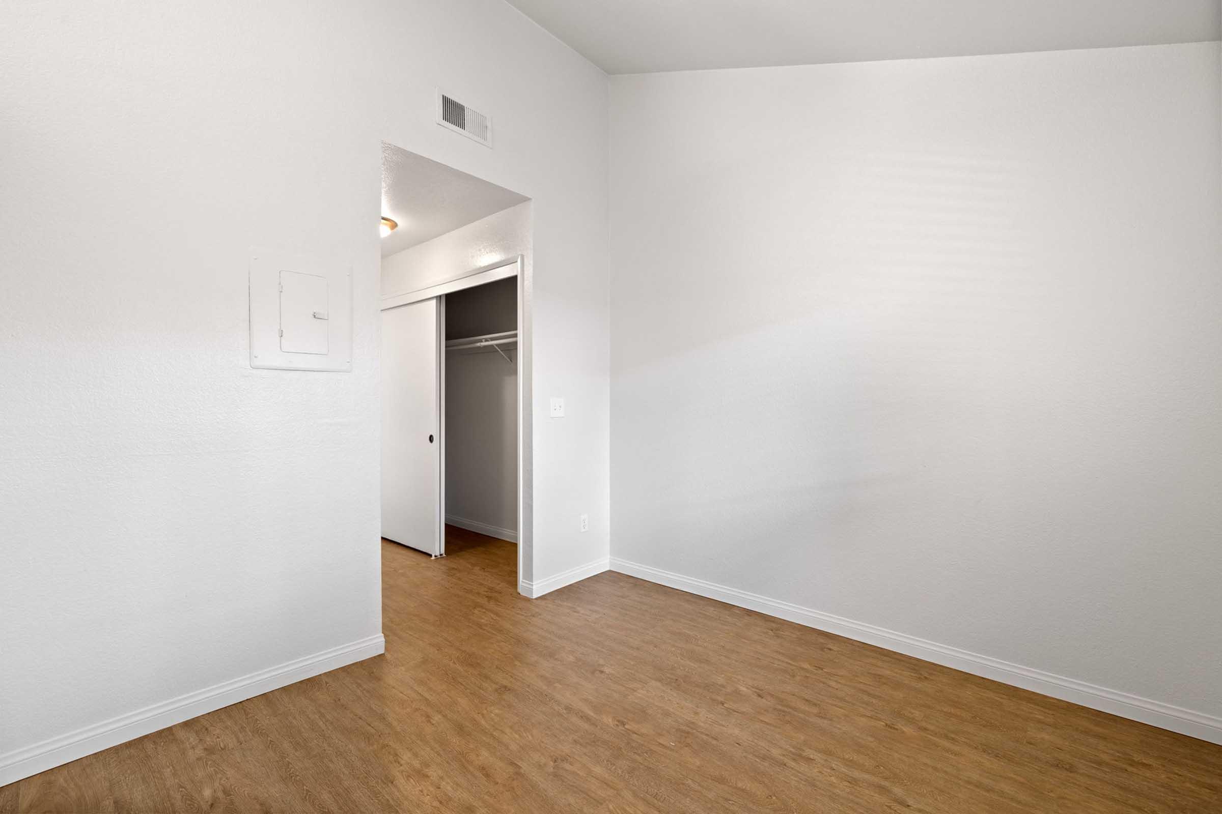 A bright, empty room with white walls and a light wood floor. There is a closet with an open door on the right side and a small recessed area near the ceiling on the left. Natural light illuminates the space, giving it a clean and airy feel.