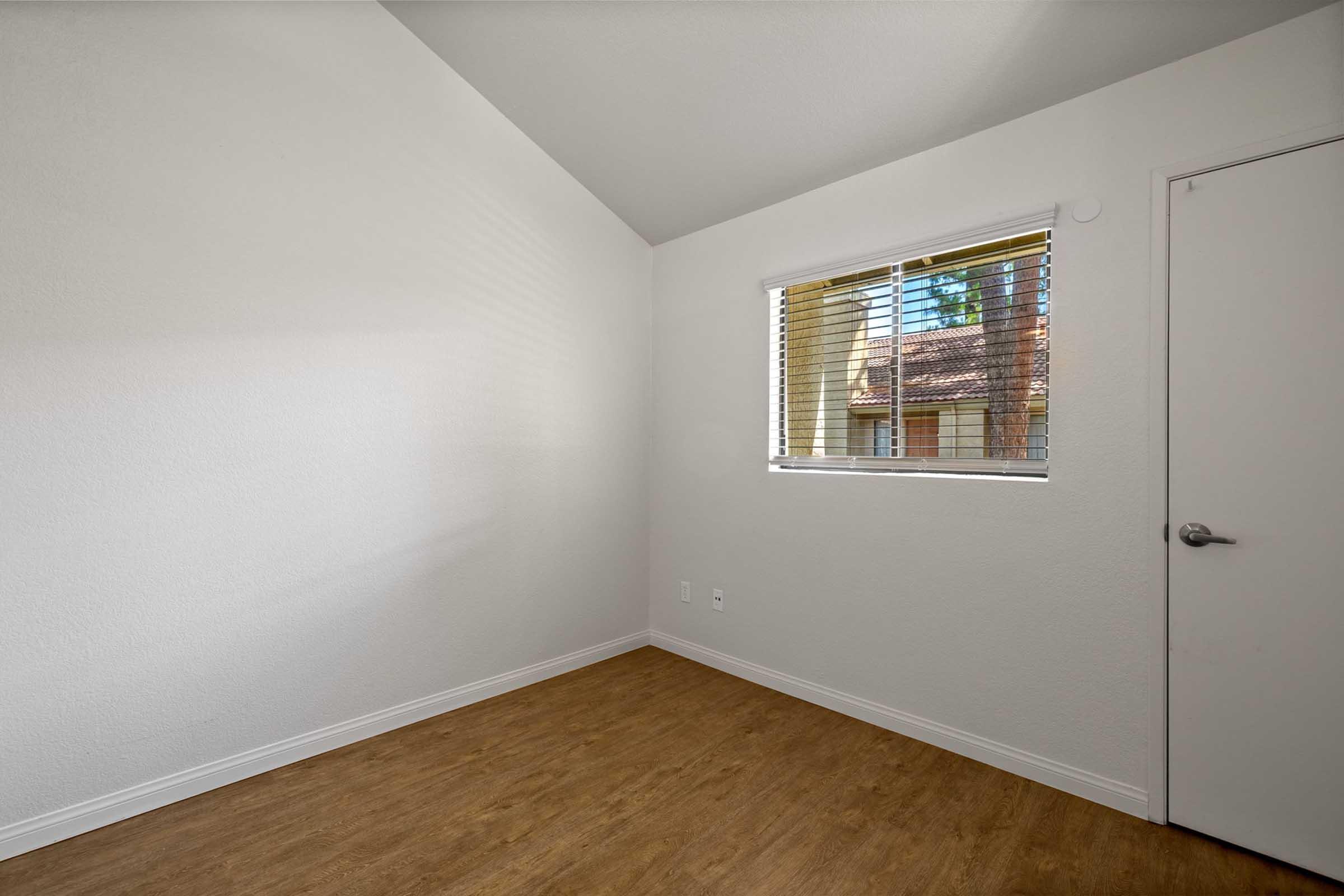 A corner of an empty room with white walls and a wooden floor. There is a window with blinds on the left side, allowing natural light to enter. The room has a high ceiling and a door visible on the right. The overall atmosphere is bright and clean, suitable for various uses.