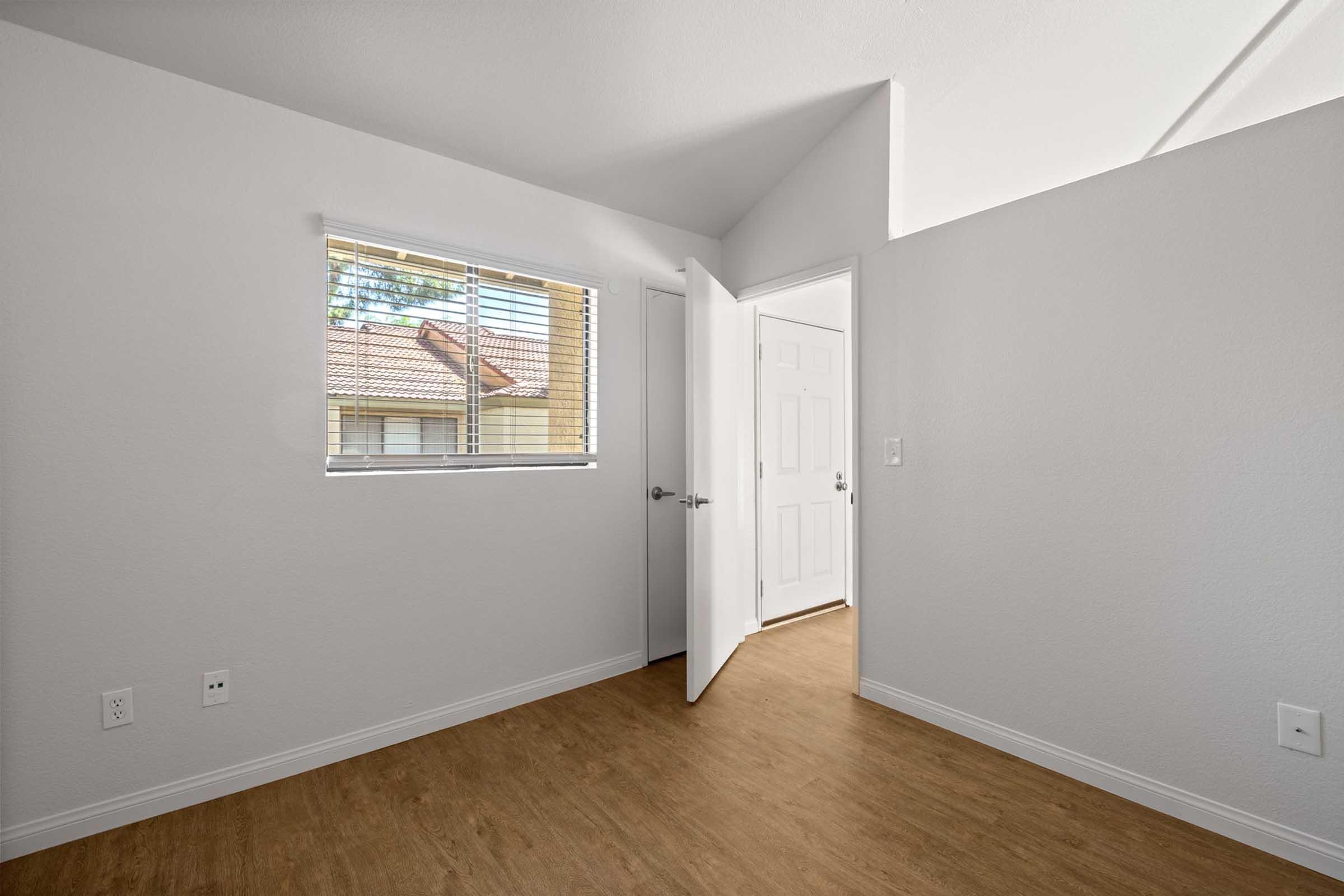 A well-lit room with light gray walls and a wooden floor. There is a window with blinds on the left, and an open door leading to another room on the right. The space appears modern and clean, with no furniture present.