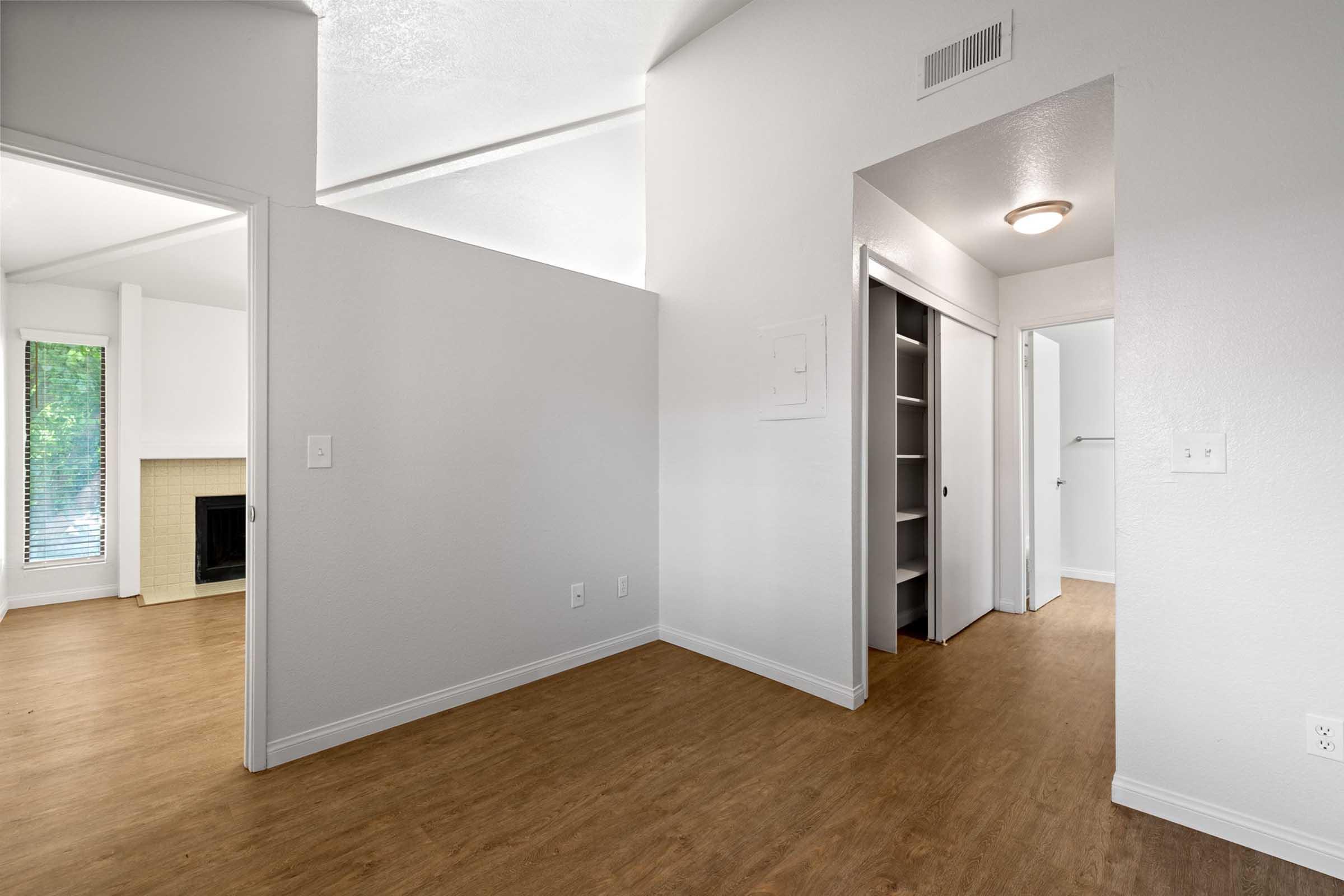 A bright, empty interior space featuring light-colored walls and wood-like flooring. On the left, there's an entryway leading to a room with a fireplace and windows. On the right, a hallway leads to a closet with sliding doors. The overall ambiance is clean and modern, suitable for various living arrangements.