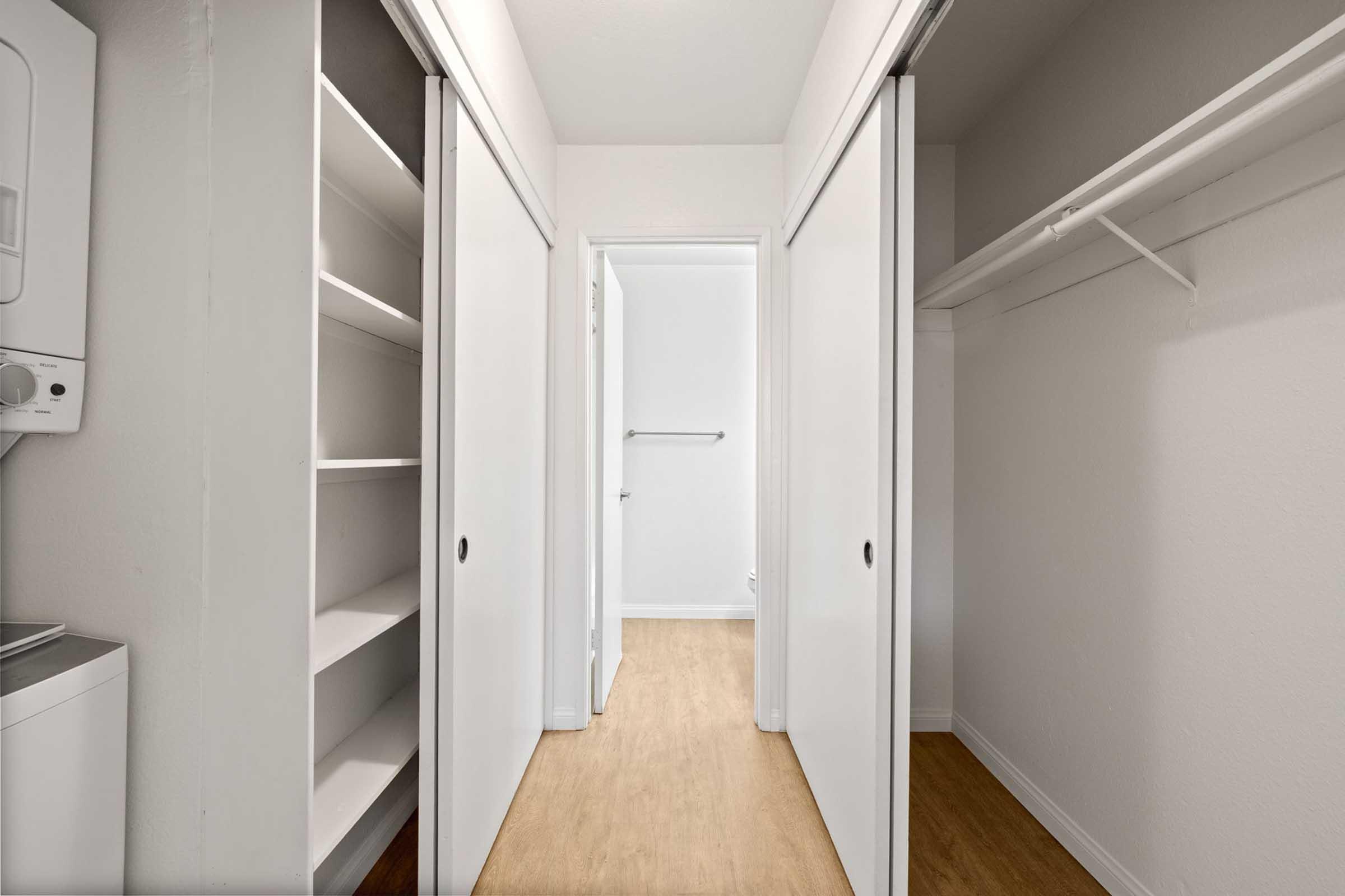 A spacious closet area with two sliding doors, featuring shelving on one side and an empty clothing rack on the other. The walls are painted a light color, and the floor has a light wood finish, creating a bright and simple storage space.