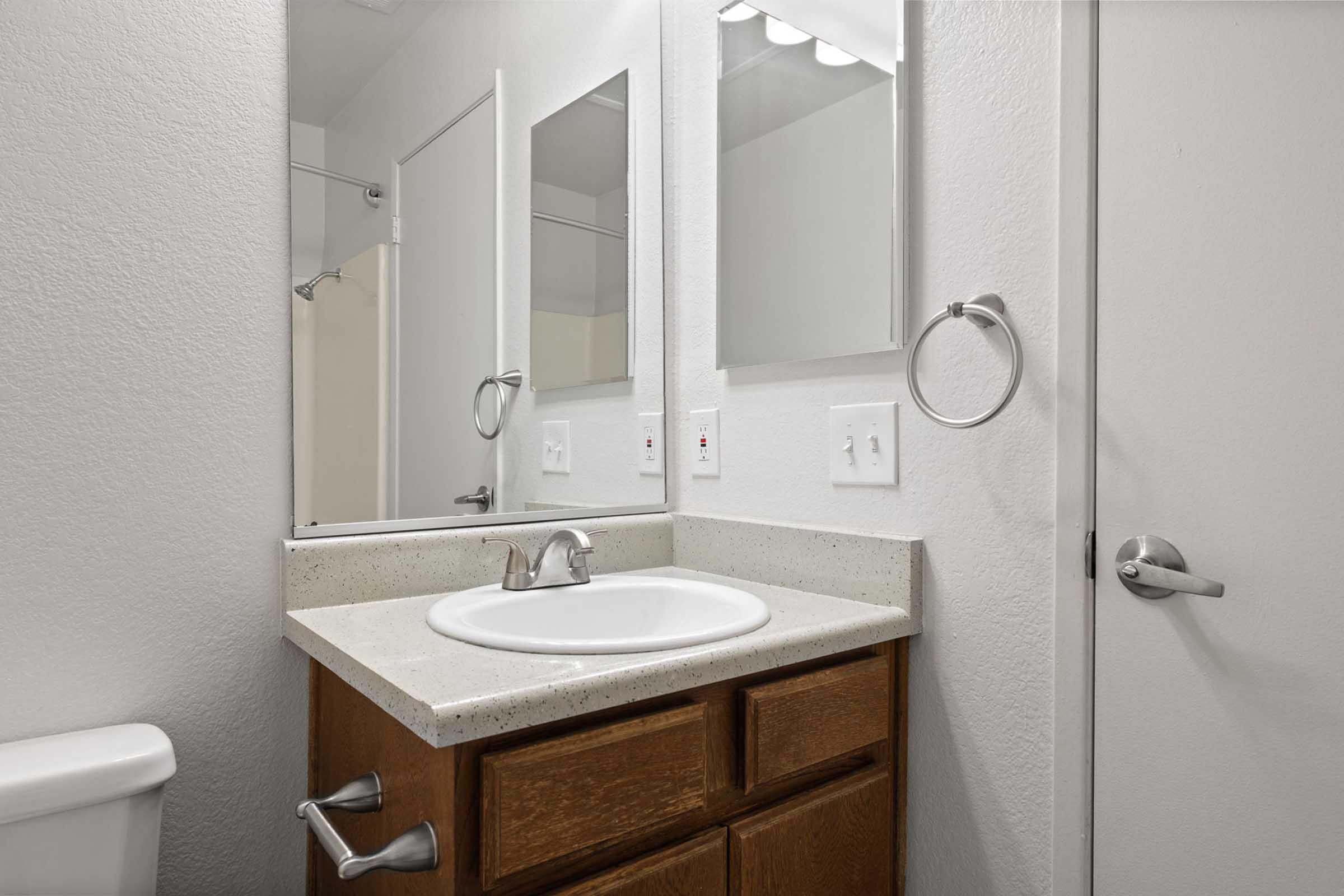 A clean, modern bathroom featuring a white countertop with a round sink, wooden cabinetry, and a wall-mounted towel holder. The wall is painted white, and there are two mirrors above the sink. A door and part of a shower area are visible in the background.