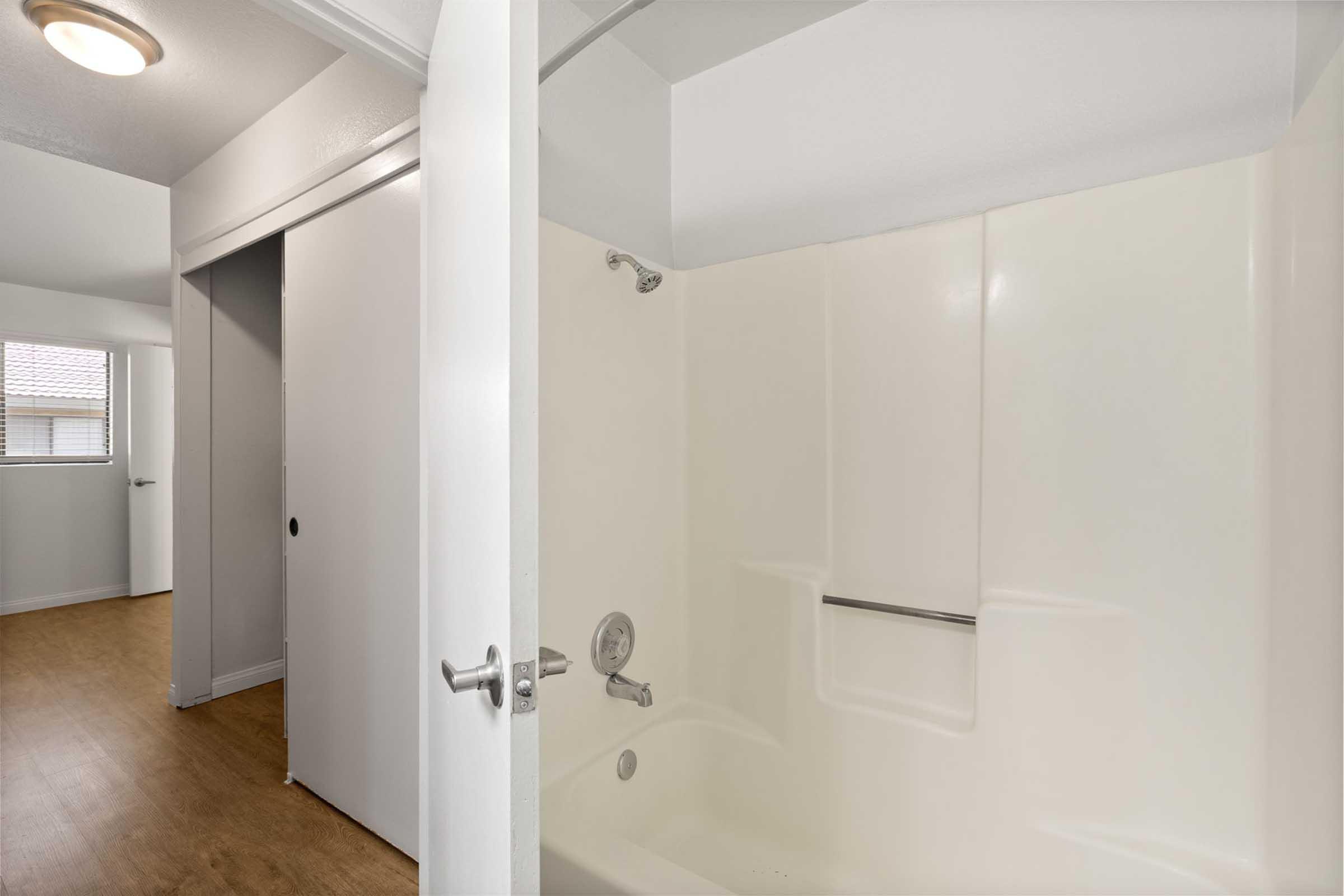 A clean, empty bathroom with a bathtub and shower fixture. The walls are a plain white, and there is natural light coming from a window outside the frame. The flooring is a light wood finish, and a closed door is visible in the background, leading to another room.