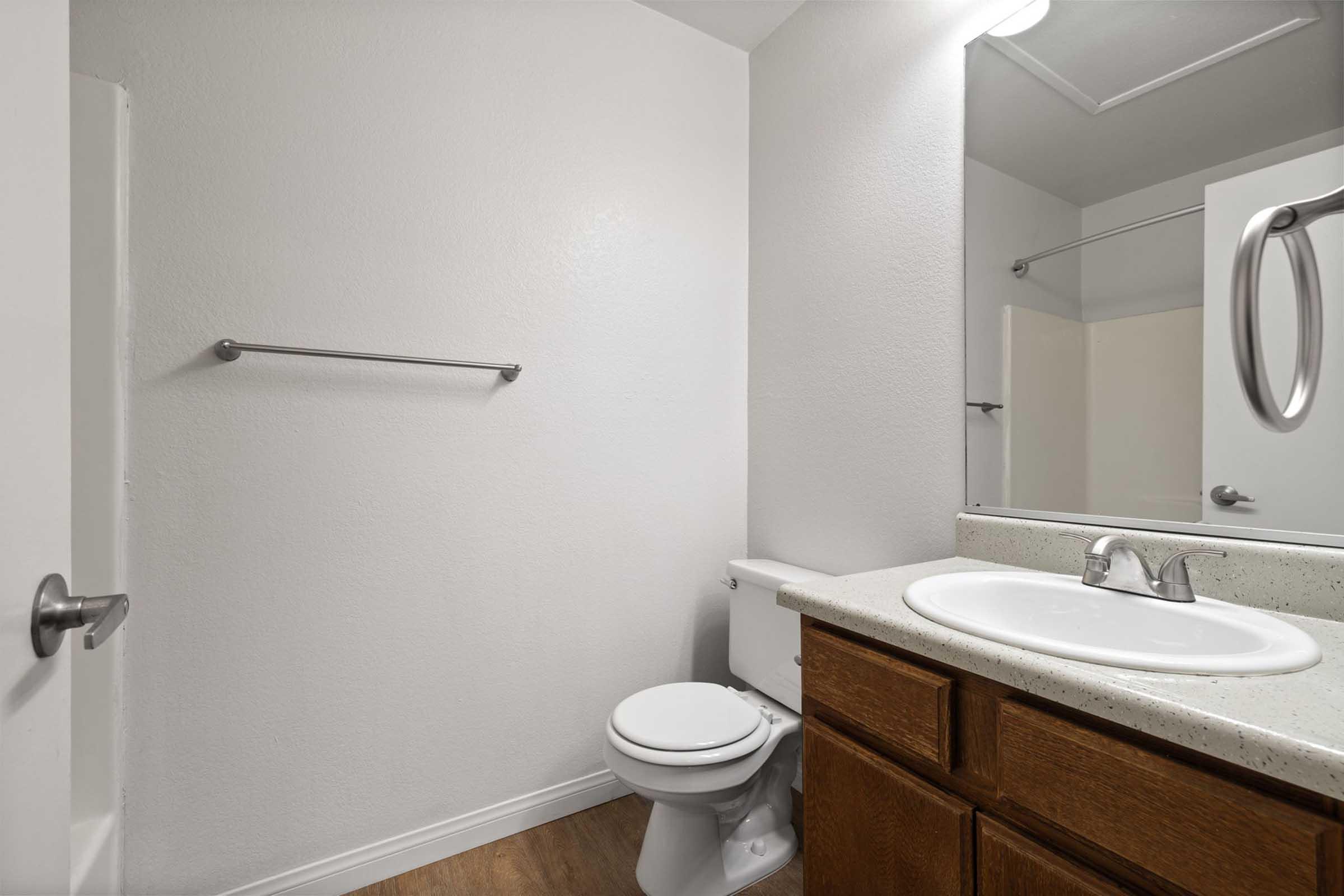 A clean and modern bathroom featuring a white wall, a wooden vanity with a round sink, a mirror above the sink, a toilet, and a towel rack. The floor is a light-colored laminate, and there is a shower area visible in the background.