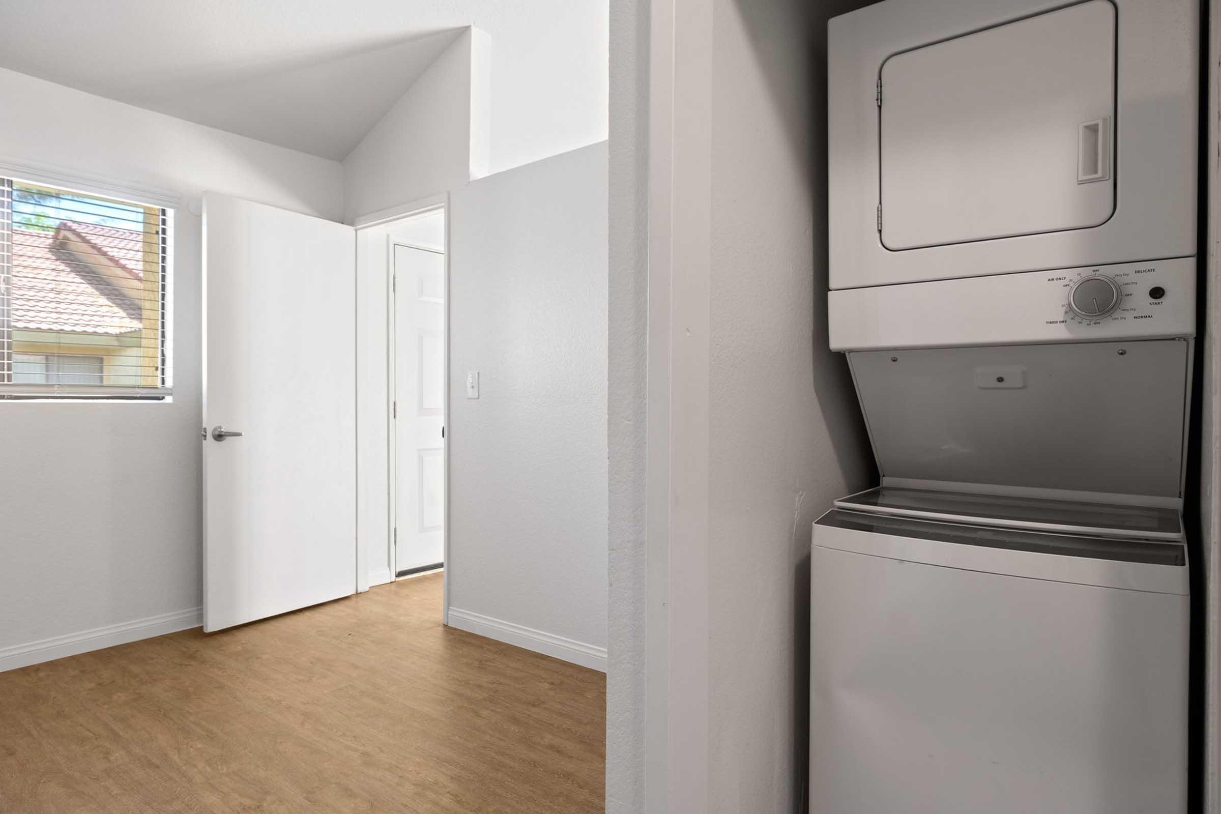A laundry area featuring a stacked washer and dryer, with a view into a bright room that has a door leading outside. Natural light enters through a window, illuminating the space with light wood flooring and neutral-colored walls.