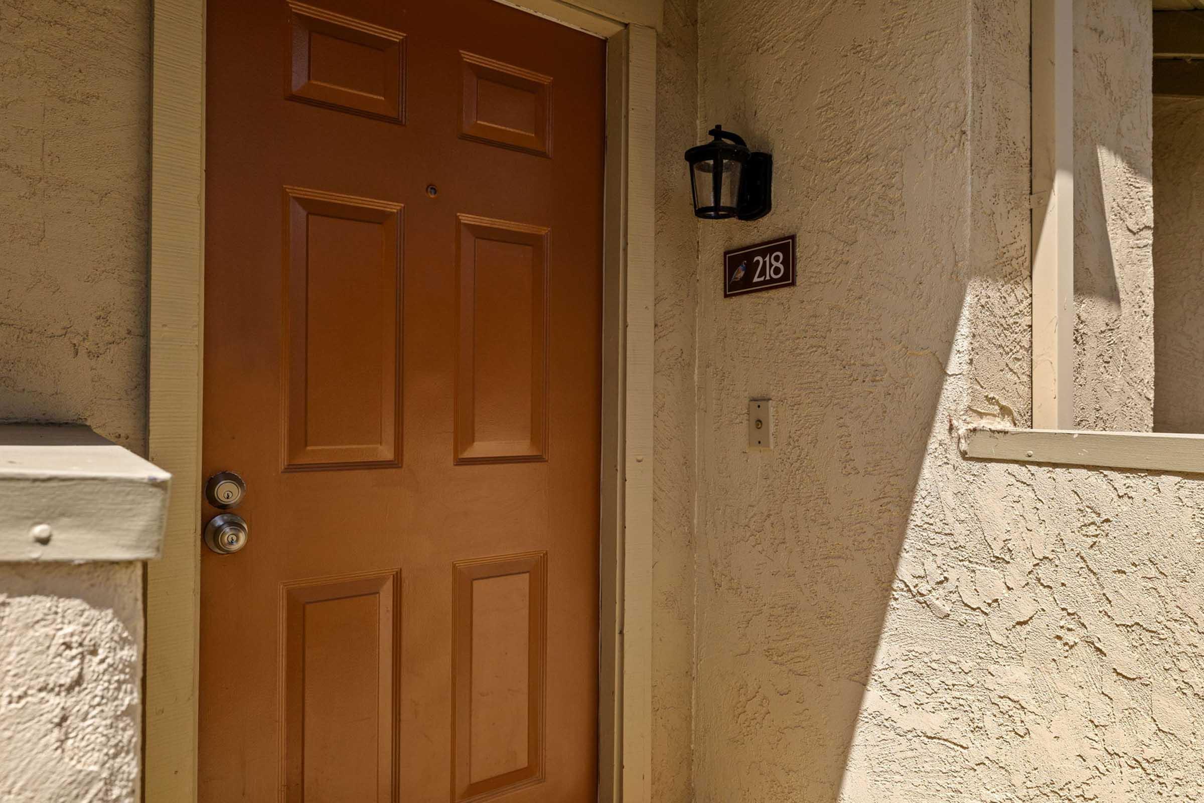 A close-up view of a brown door with a silver doorknob, located at room number 218. The door is set into a textured beige wall, and there is a black lantern-style light fixture mounted beside it. The overall scene presents an inviting entrance to a residential or commercial space.