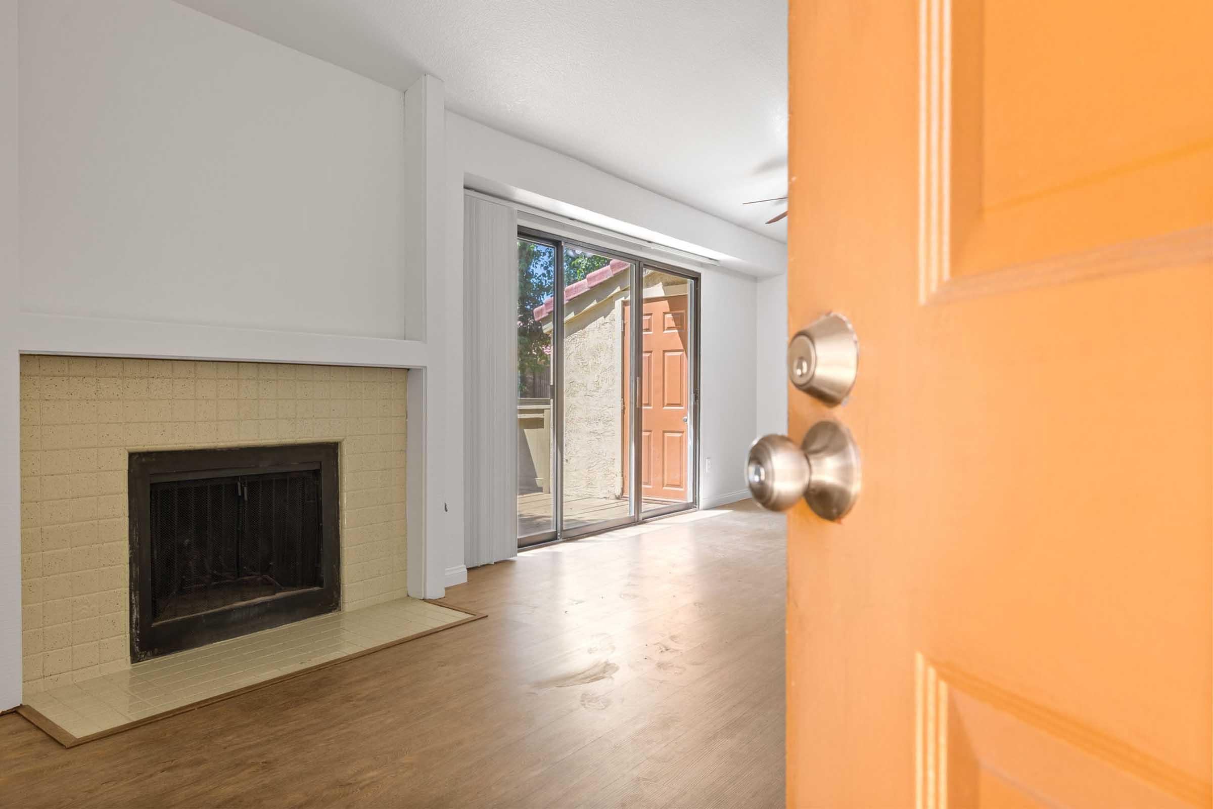 View from an open orange door into a cozy living room featuring a brick fireplace and large sliding glass doors that lead to an outdoor area. The space has a warm wooden floor and neutral walls, creating an inviting atmosphere. Natural light brightens the room.