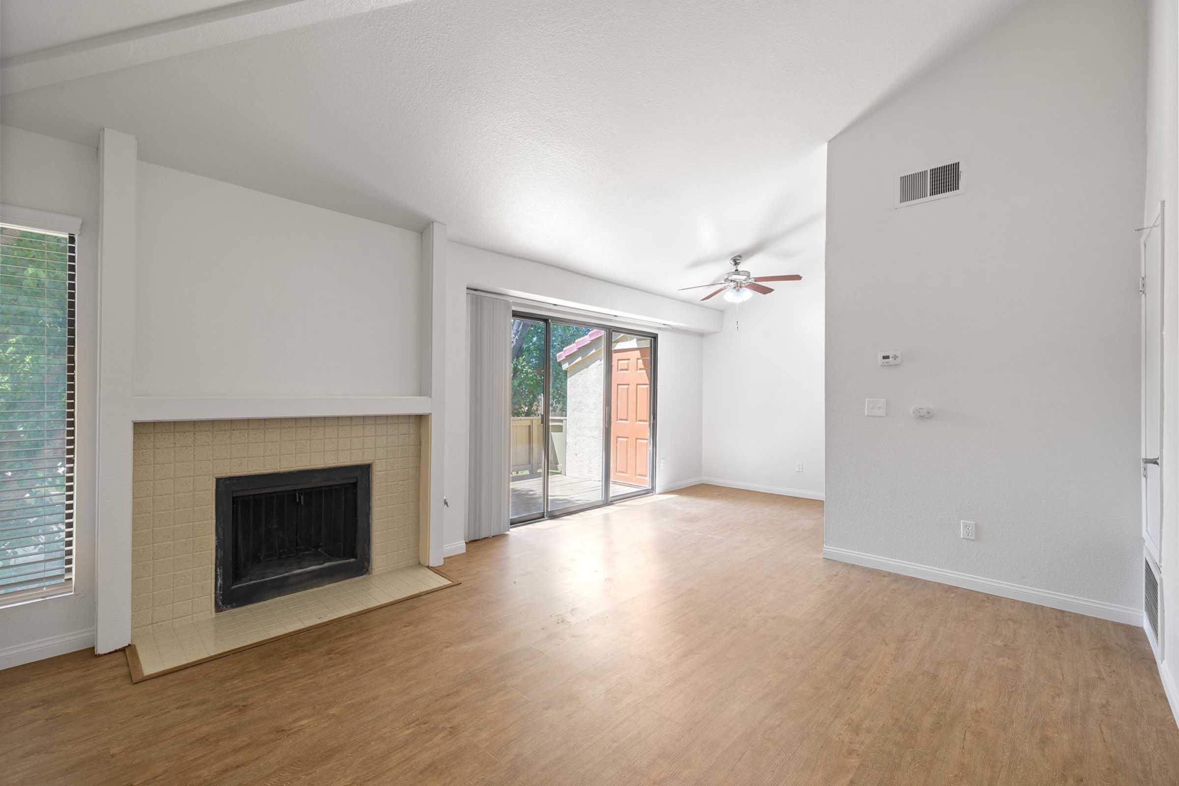 A bright and open living room featuring a fireplace, large windows allowing natural light, and access to an outdoor space through glass sliding doors. The walls are painted in a light color, and the flooring is a warm wood-like laminate. Ceiling fan is installed above.