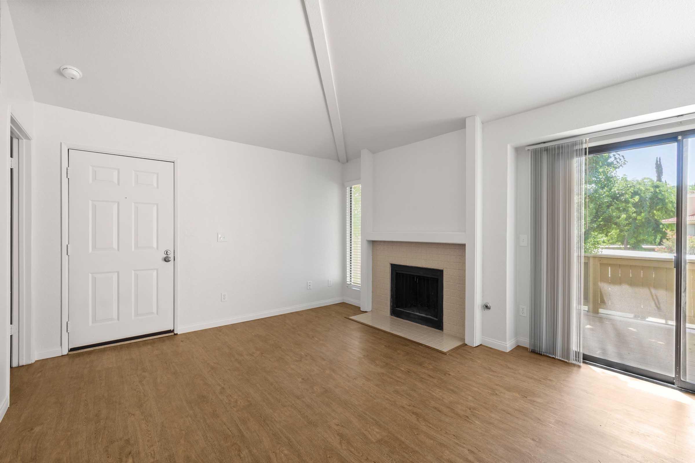 Spacious living area featuring a large window and sliding glass door that opens to a balcony. A fireplace is located against the wall, and the room has light-colored walls and a wooden floor. The ceiling is high with exposed beams, creating an open and airy atmosphere.