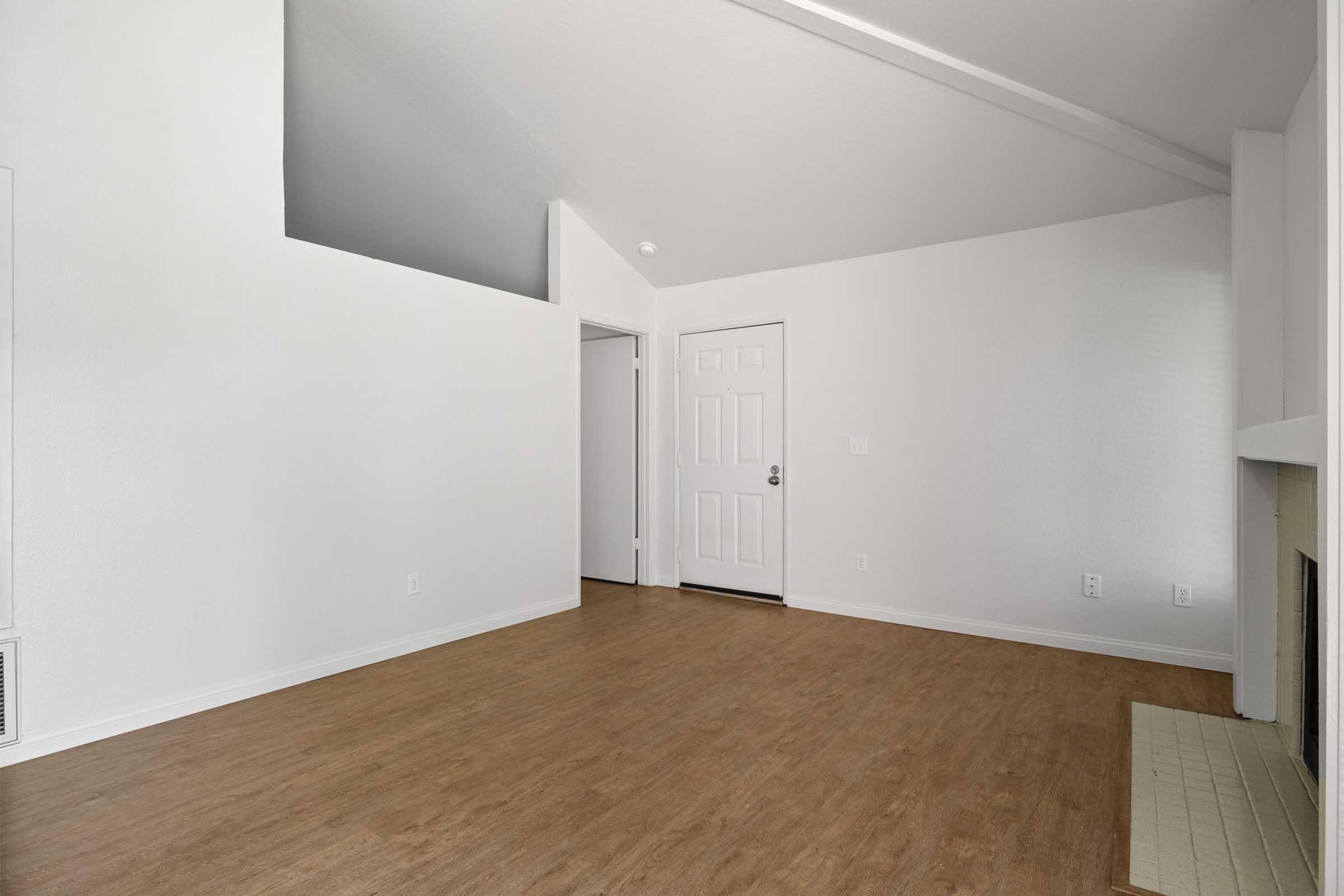 A spacious, empty living room featuring light-colored walls and a large window. The floor is covered with light brown laminate. There is a doorway leading to another room, and a fireplace on one side of the room. The ceiling has a slight slope, enhancing the room's openness.