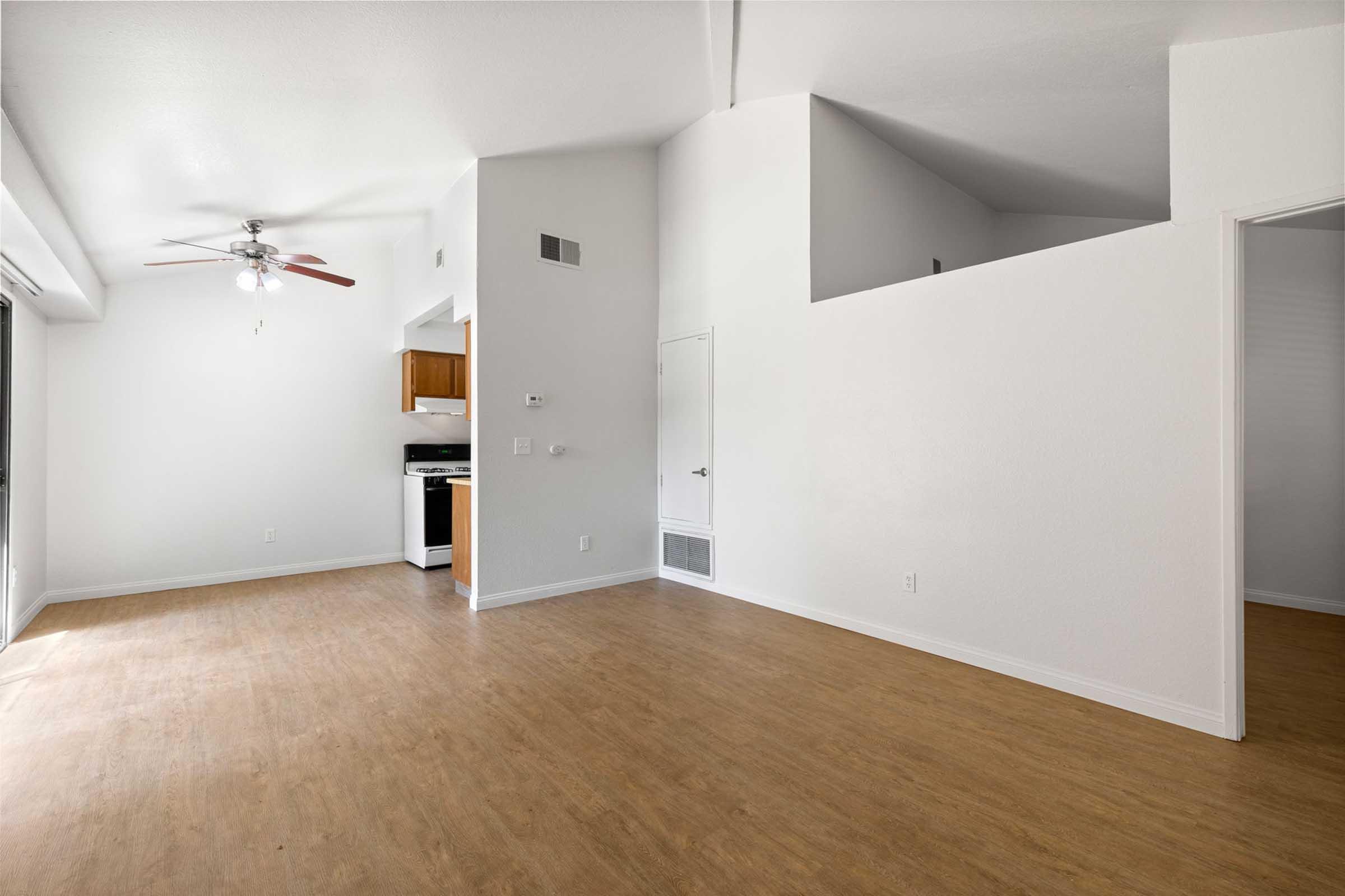 A spacious, empty living room with light-colored walls and wood-like flooring. A ceiling fan is mounted, and there is a small kitchen area visible with cabinets and a stove. A doorway leads to another room, and natural light fills the space from a window.