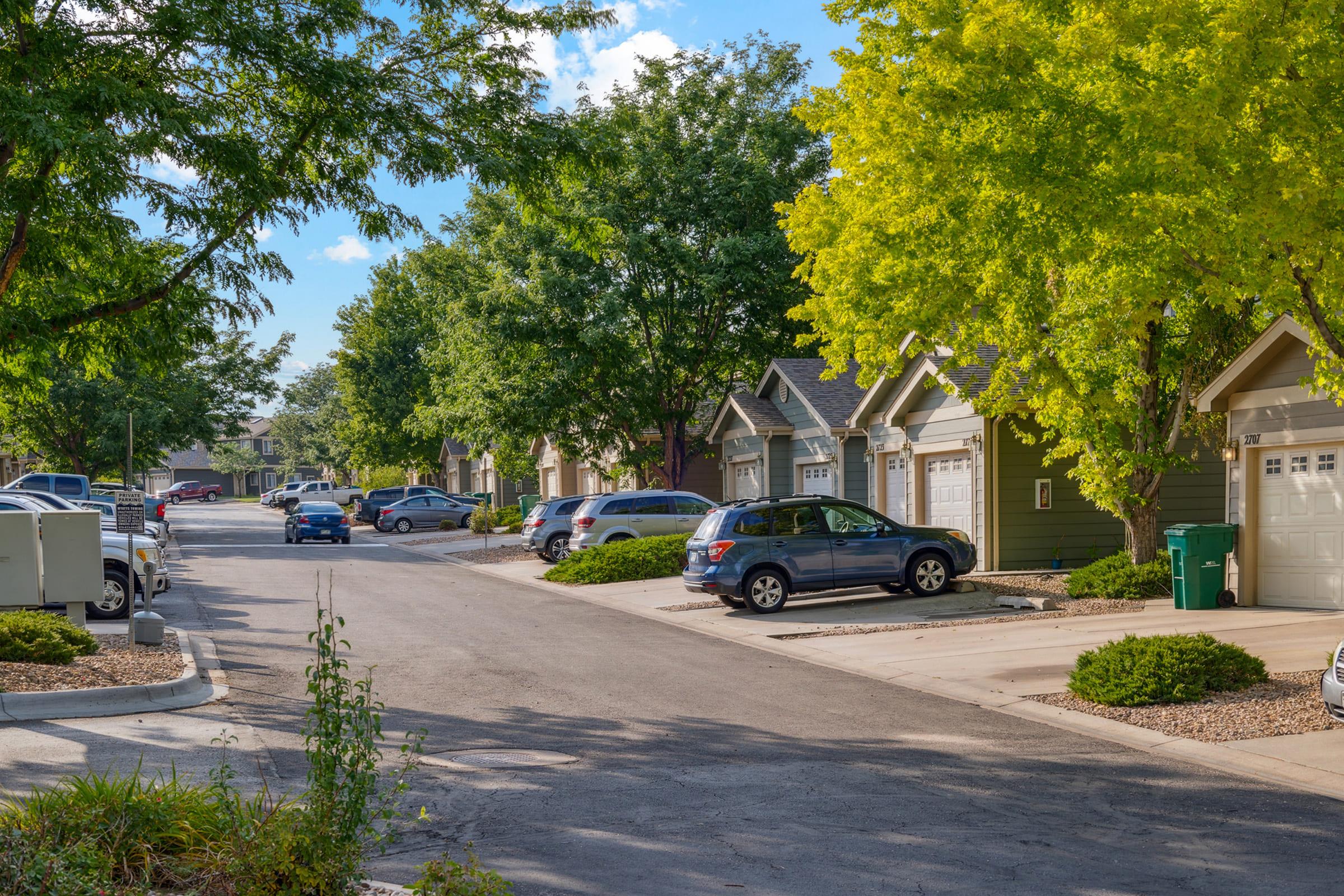 A quiet residential street lined with green trees and houses. Several parked cars are visible along the curb, and well-maintained lawns and shrubs add to the neighborhood’s charm. The sky is clear with a few clouds, creating a pleasant atmosphere.