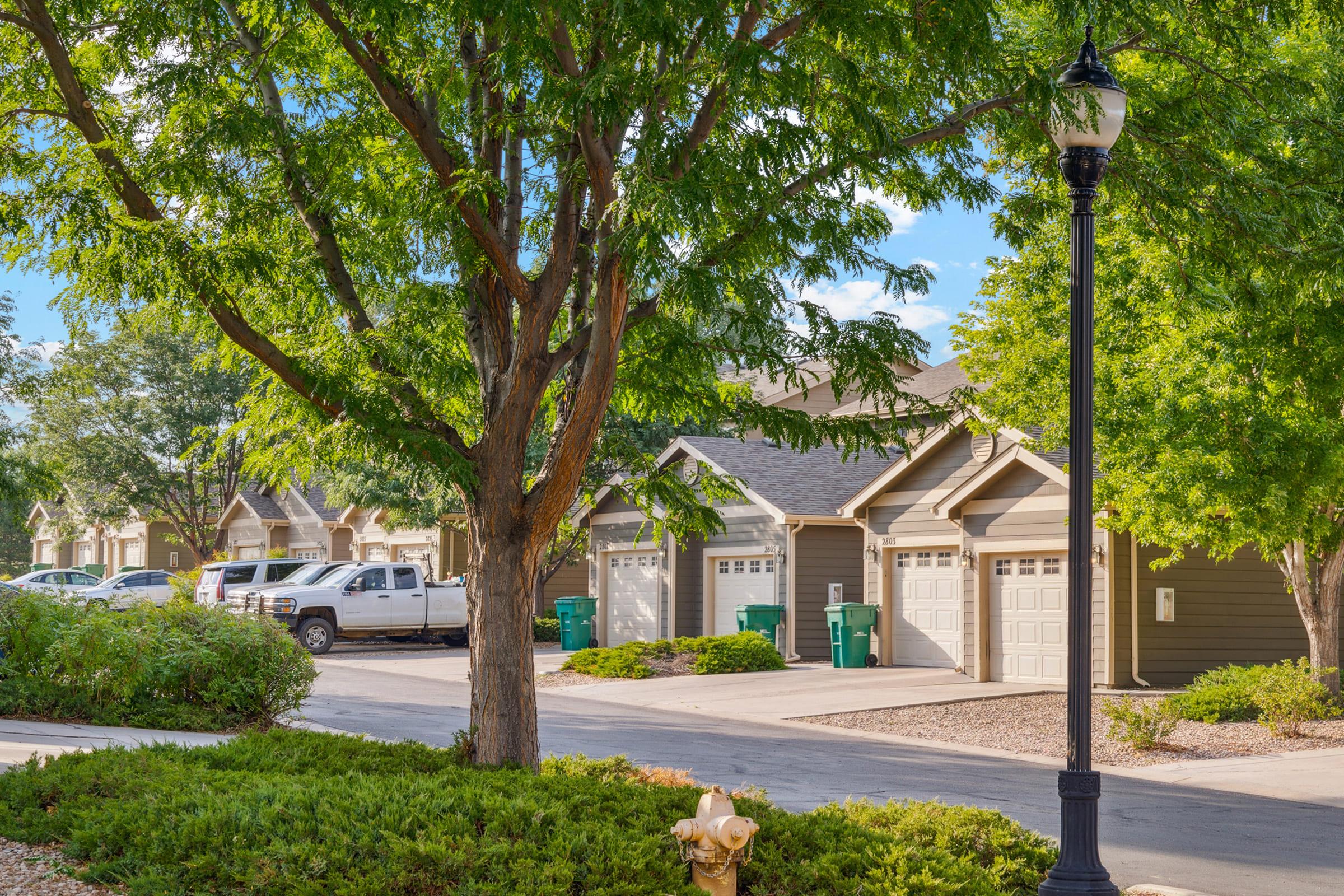 A residential neighborhood featuring several houses with garages, a tree-lined street, and a lamppost. Green trash bins are placed near the garages, with a white pickup truck parked in the driveway. Lush greenery surrounds the area, creating a peaceful suburban atmosphere.