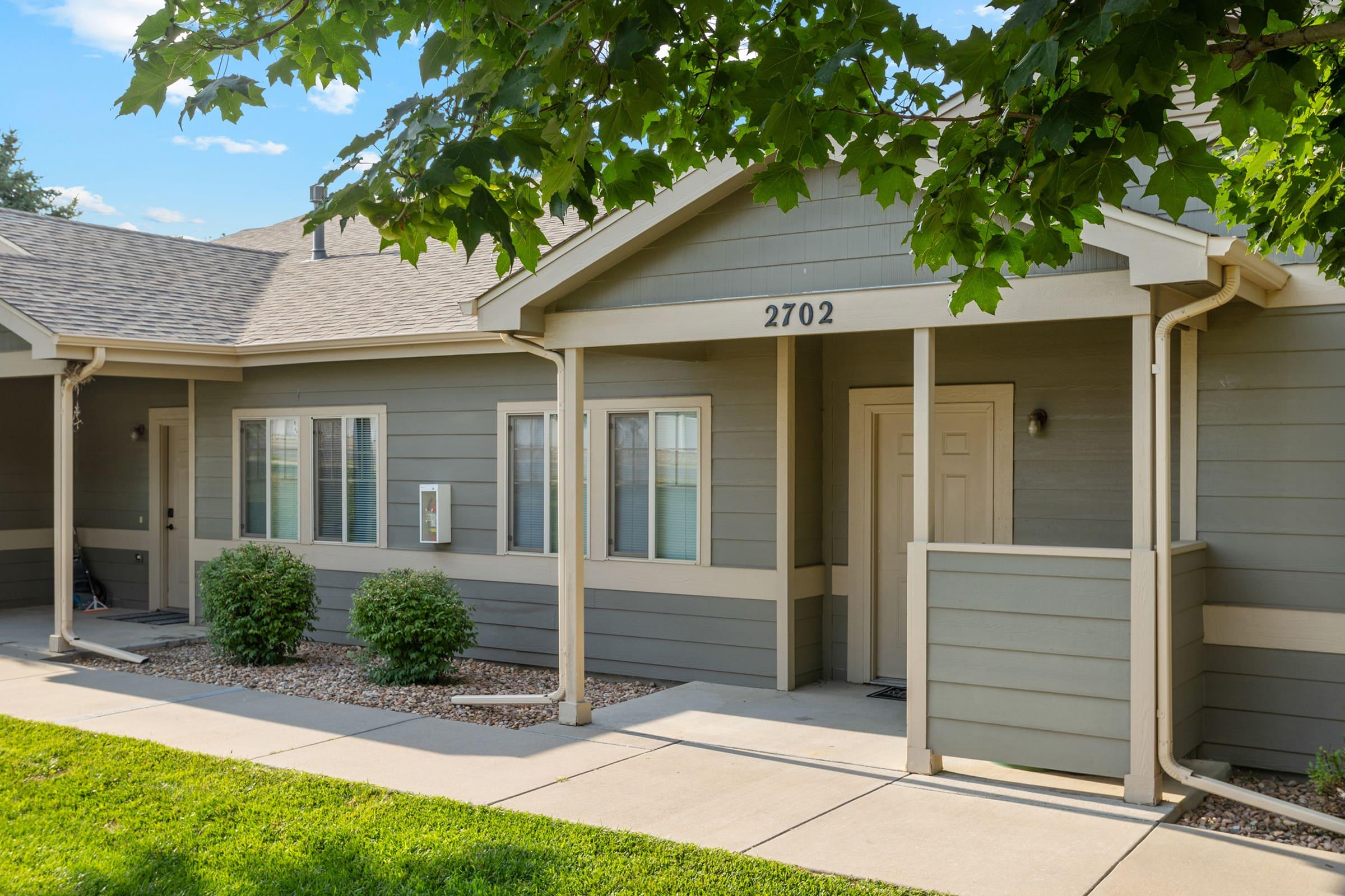 A modern, single-story residential building with grey siding and a welcoming front porch. The address "2702" is displayed above the door. The entryway features neatly trimmed bushes and a manicured lawn, with a pathway leading to the entrance under a clear blue sky.