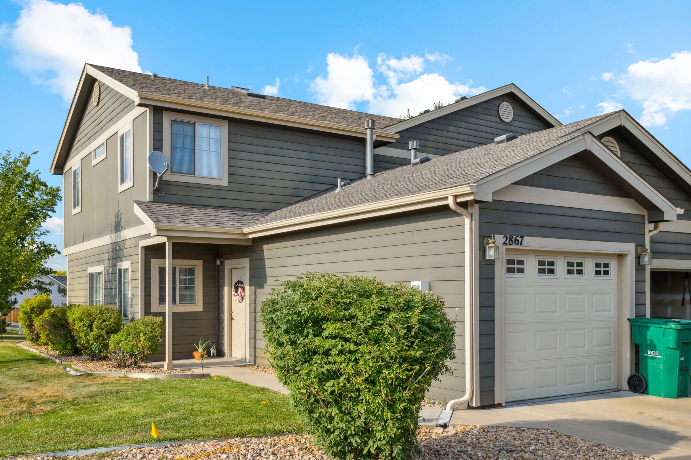 A modern two-story townhouse with gray siding and a garage. The front yard features a small lawn and neatly trimmed shrubs. There are two recycling bins on the side. The sky is clear with a few clouds, and the overall setting appears well-maintained and residential.
