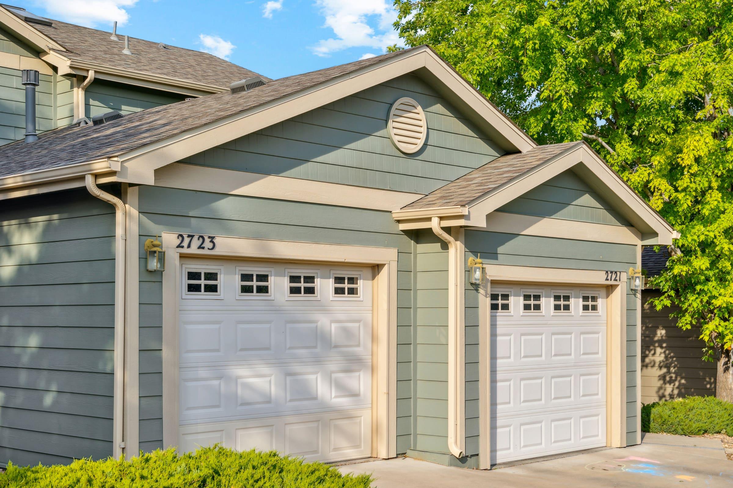 Two adjacent garage doors on a residential property, one labeled "2723." The building features a light green exterior with white trim and a decorative vent above the doors. Surrounding vegetation includes lush green trees, and the sky is partly cloudy.