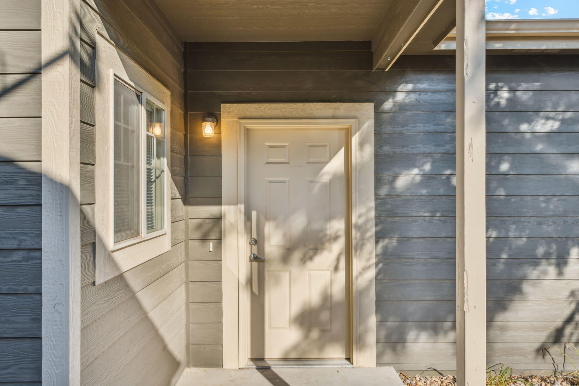 A close-up view of an entrance featuring a beige door with a simple handle, framed by gray wooden siding. A window with white shutters is visible to the left, and a light fixture is mounted above the door. Soft shadows fall across the entrance area, enhancing a welcoming atmosphere.