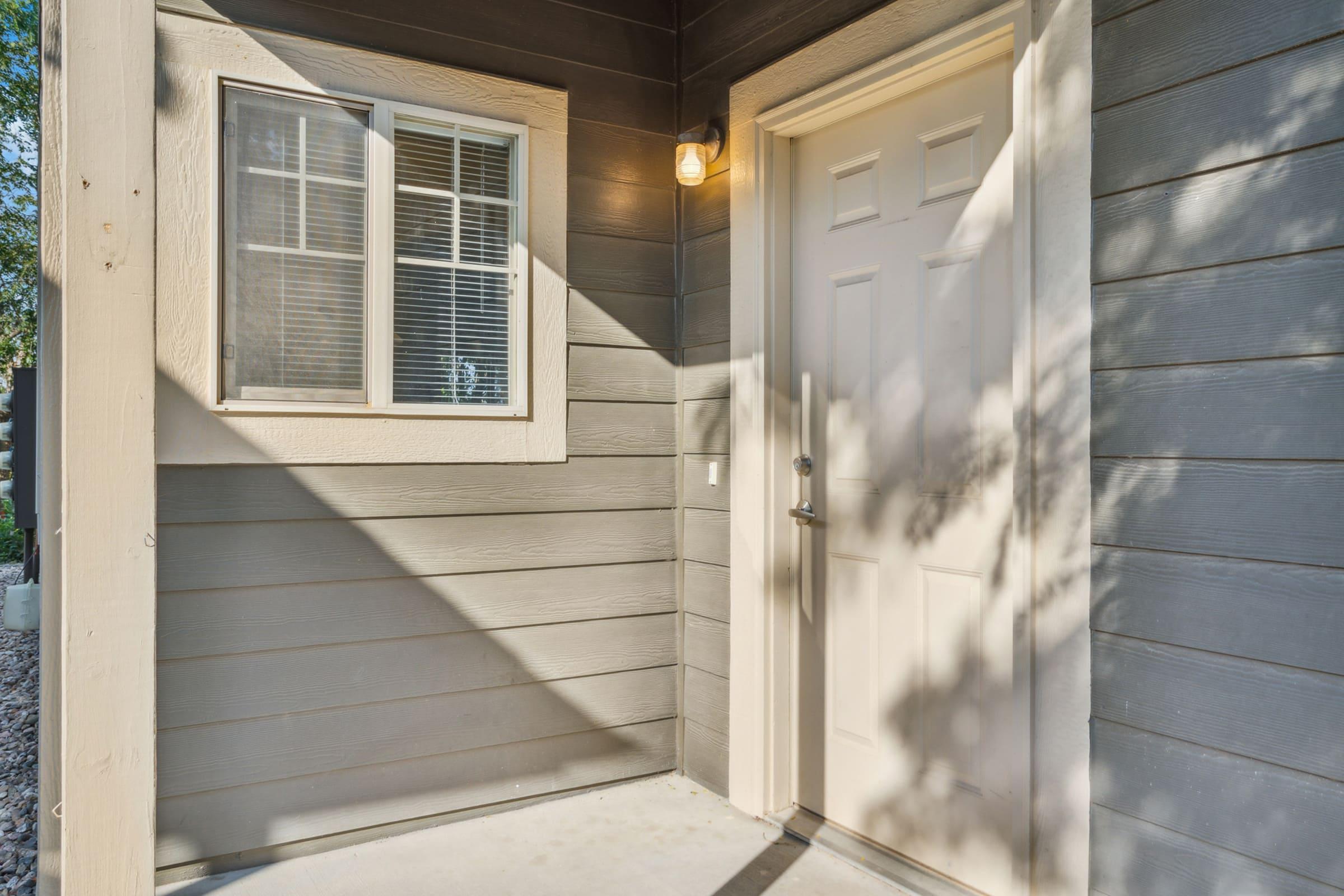 A close-up view of a home's entrance featuring a light fixture above a closed door with a doorknob, framed by a wooden wall with horizontal siding. A window is visible beside the door, casting shadows from nearby foliage. The scene is well-lit, suggesting a bright day.