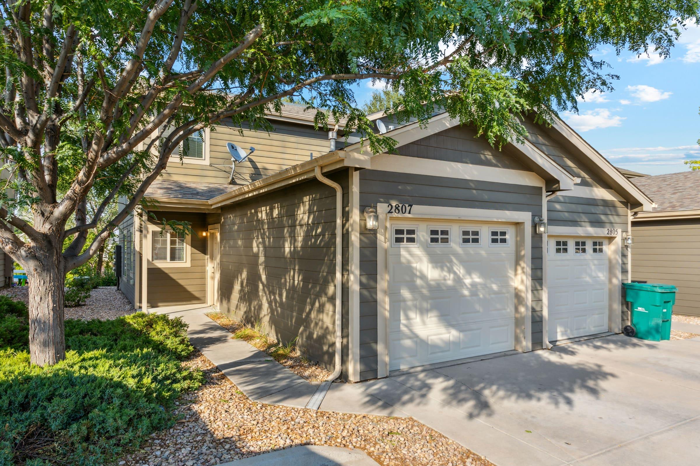 A modern two-story house with a light brown exterior, featuring a double garage and a walkway surrounded by landscaped bushes and trees. A green trash bin is visible to the left. The home is well-lit, with clear blue skies and some clouds in the background.