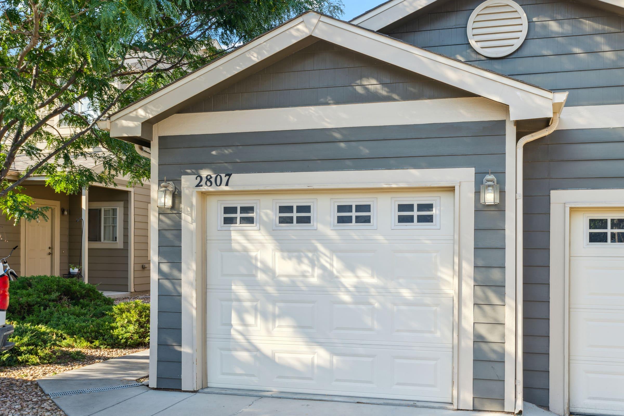 A residential home's exterior featuring a garage door numbered 2807, painted in light gray. The garage has rectangular windows at the top, flanked by two light fixtures. Surrounding the house are trees and landscaped greenery.
