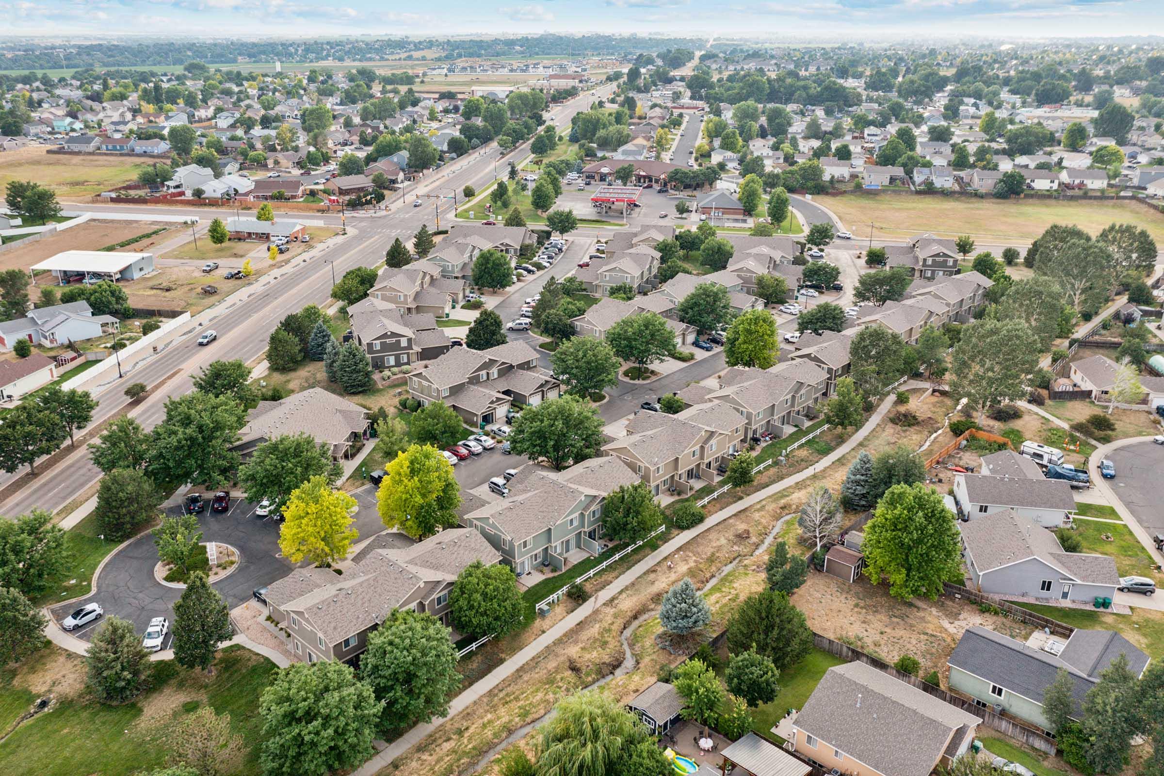 Aerial view of a suburban neighborhood featuring numerous houses with grey roofs, tree-lined streets, and green patches. The layout shows a mix of residential areas and nearby roads, with some open fields visible in the background. The scene conveys a peaceful, community-oriented atmosphere.