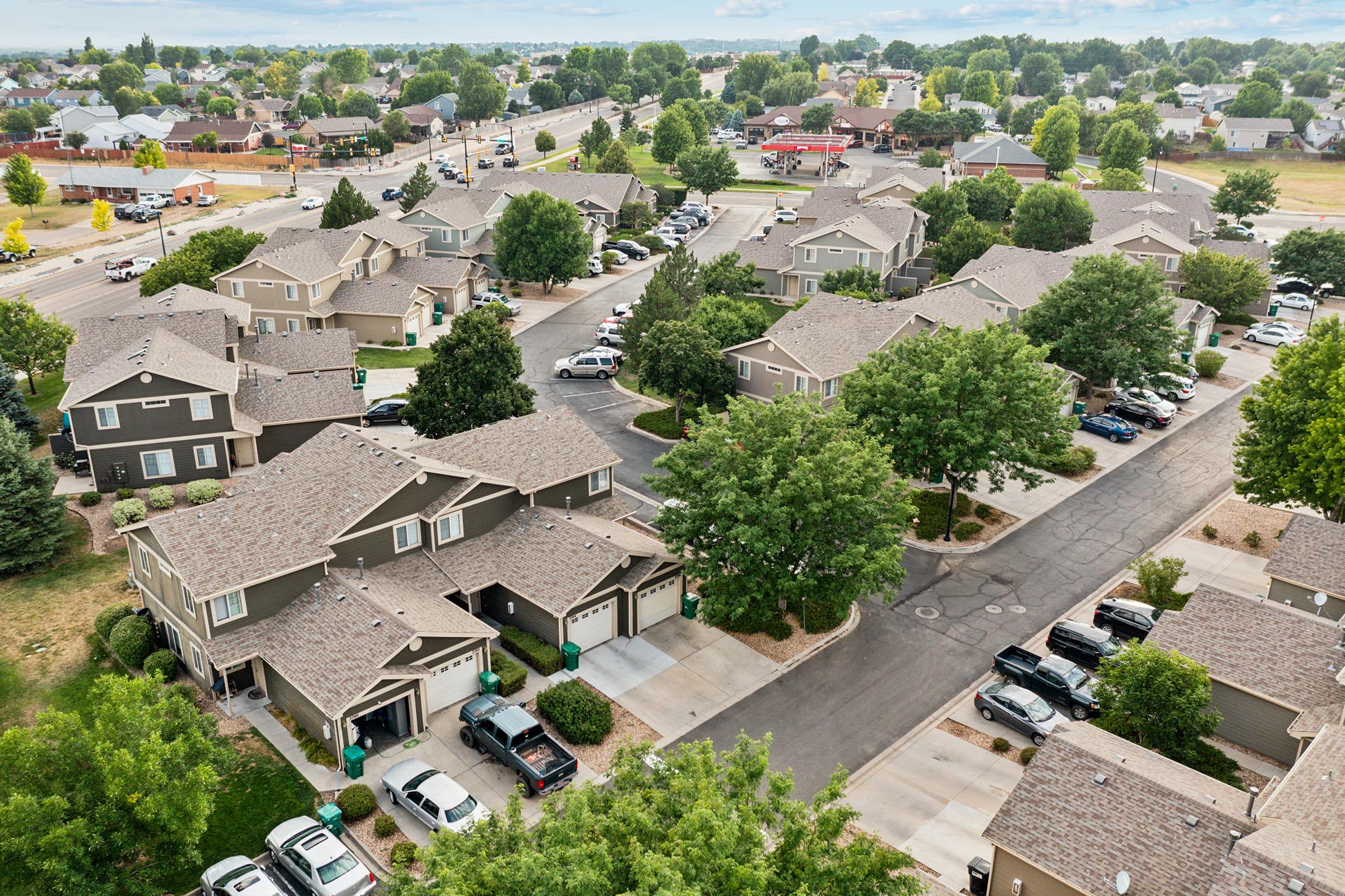 An aerial view of a suburban neighborhood featuring several houses with similar architecture. The streets are lined with trees, and vehicles are parked in driveways and along the road. The scene captures a peaceful residential atmosphere with a mix of greenery and well-maintained properties.