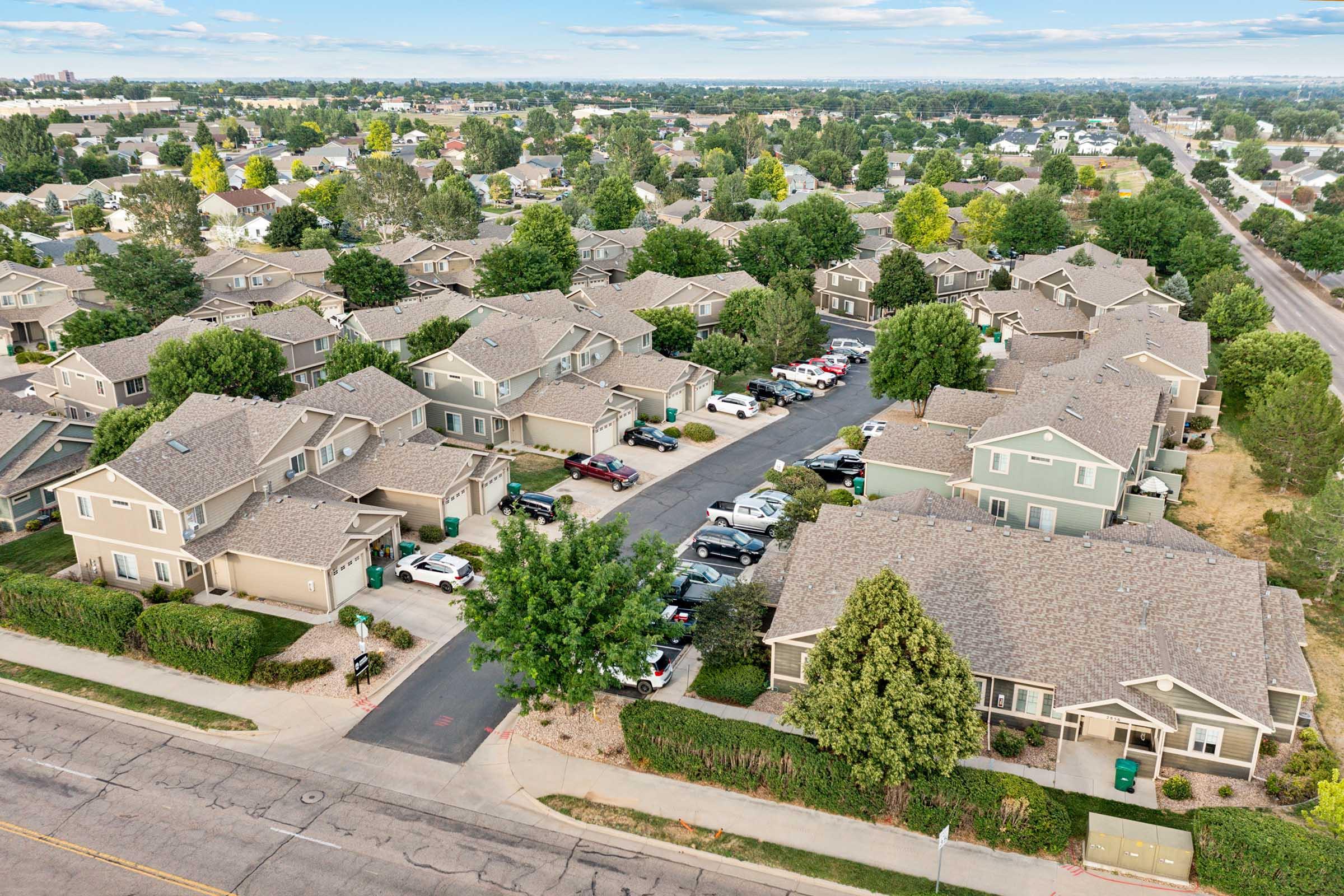Aerial view of a suburban neighborhood featuring multiple houses with gray roofs, tree-lined streets, and parked cars along the roads. The landscape includes green lawns and patches of trees, with a mix of residential properties and open spaces. In the distance, more houses and a blue sky are visible.