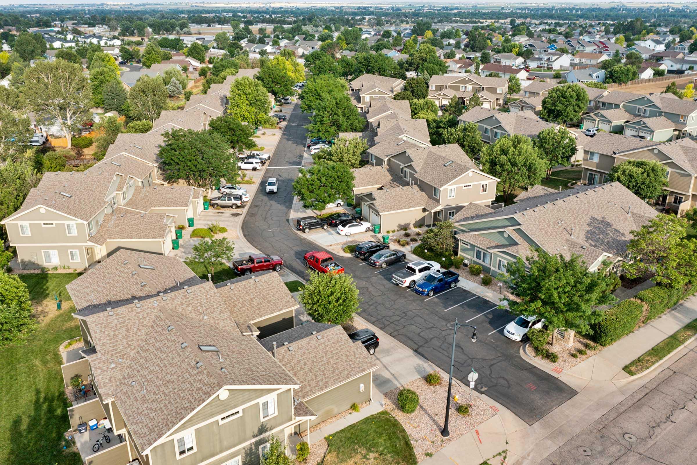 Aerial view of a suburban neighborhood featuring rows of single-family homes with sloped roofs, tree-lined streets, and parked cars. Lush green lawns and landscaped yards add to the residential setting, with a clear blue sky in the background. The layout shows a quiet, family-friendly community.
