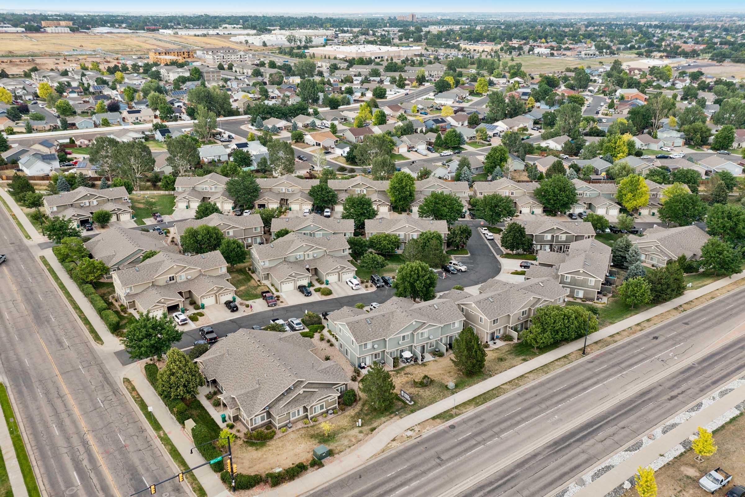 Aerial view of a suburban neighborhood featuring numerous residential homes, tree-lined streets, and parked cars. A main road runs along the bottom of the image, with additional developments visible in the background, indicating a well-established community surrounded by open land.