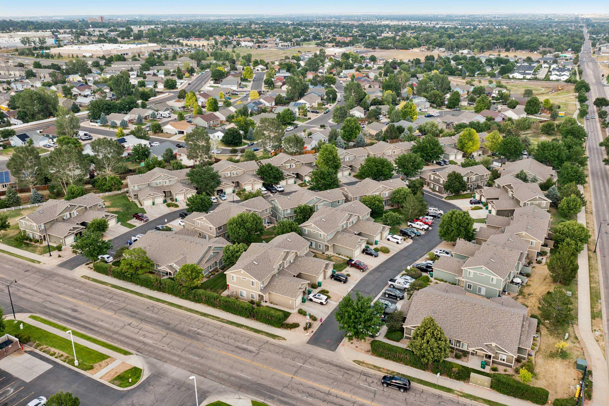 Aerial view of a suburban neighborhood featuring several residential buildings with sloped roofs, tree-lined streets, and parked cars. In the background, more houses and open spaces are visible, indicating a well-planned community. Clear skies above suggest a sunny day.