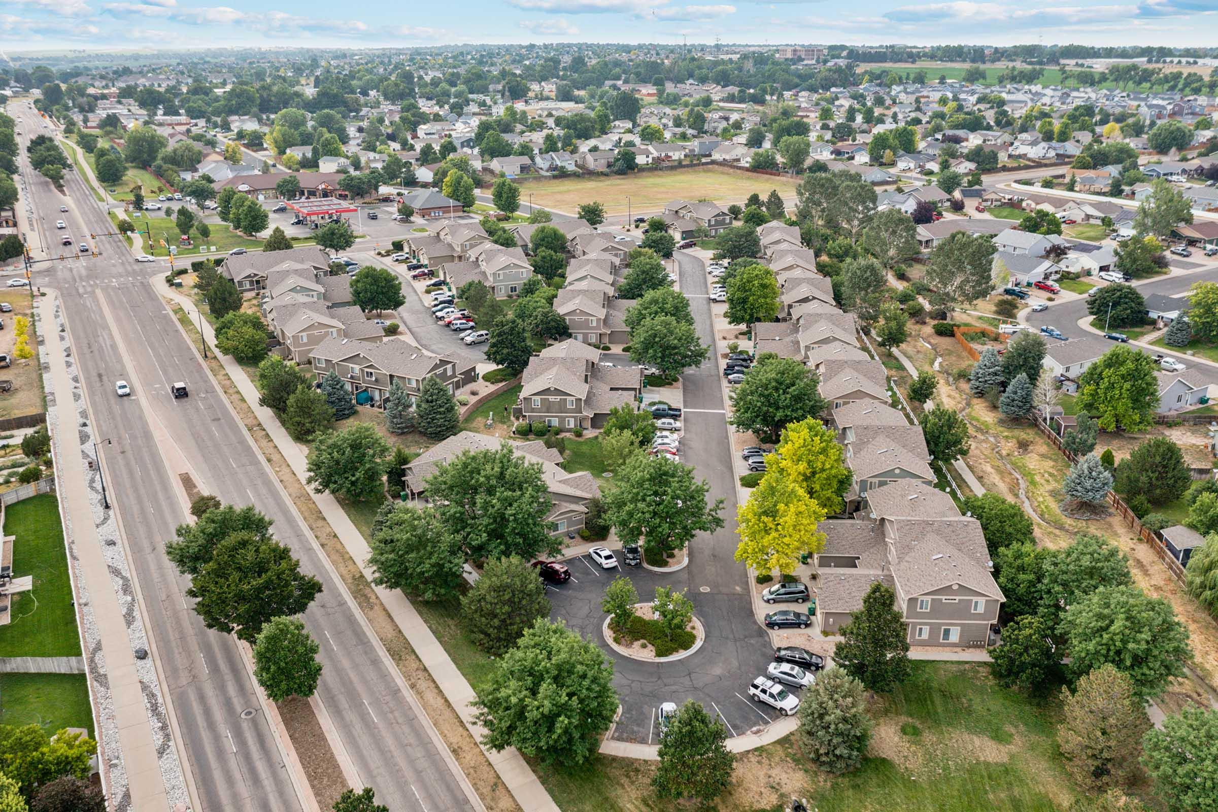Aerial view of a suburban neighborhood featuring several apartment complexes arranged in a circular layout, surrounded by trees. In the background, a wider view of the town with roads and greenery is visible, showcasing residential areas and fields.