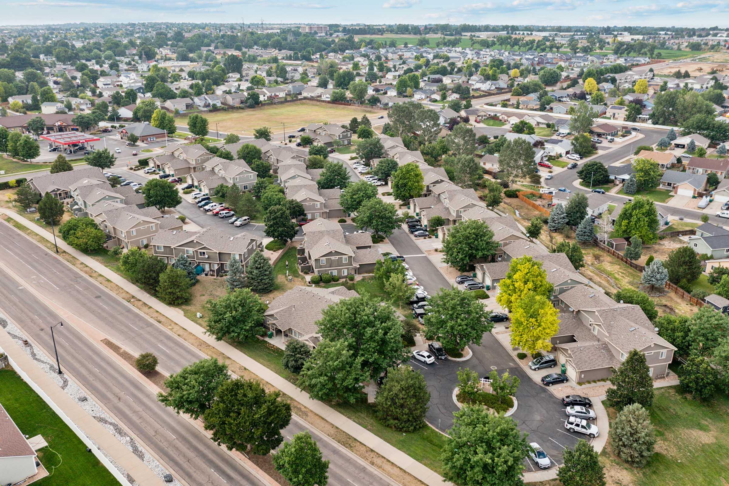 Aerial view of a residential neighborhood featuring clusters of homes, green trees, and parked cars. A road runs alongside the area, with additional buildings and open spaces visible in the distance. The landscape shows a mix of developed and natural areas, typical of suburban living.