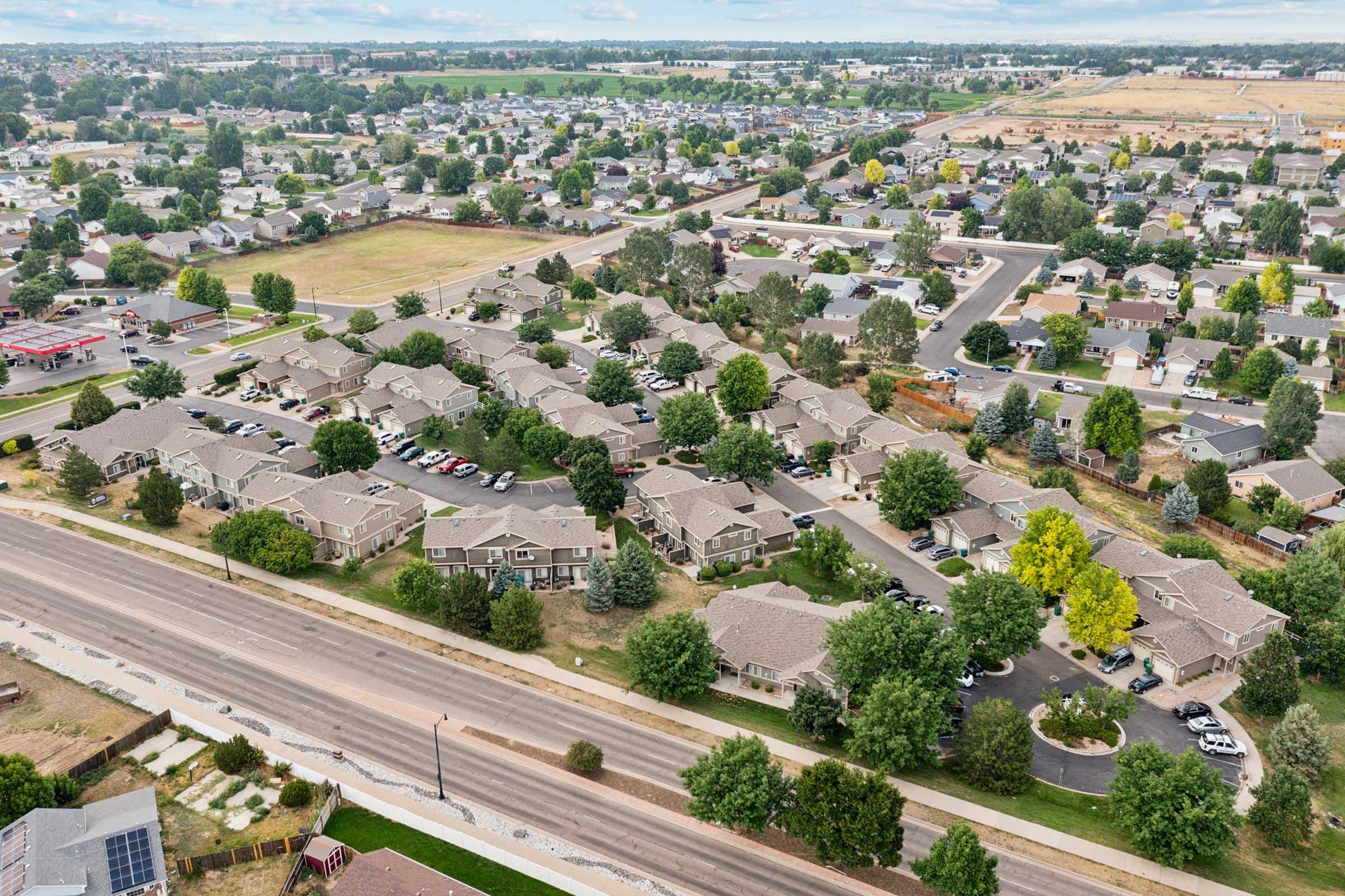 Aerial view of a suburban neighborhood featuring clusters of homes, green trees, and well-maintained roads. The landscape shows a mix of residential areas and open fields, with parked cars visible along the streets. In the distance, more houses and open spaces can be seen under a partly cloudy sky.
