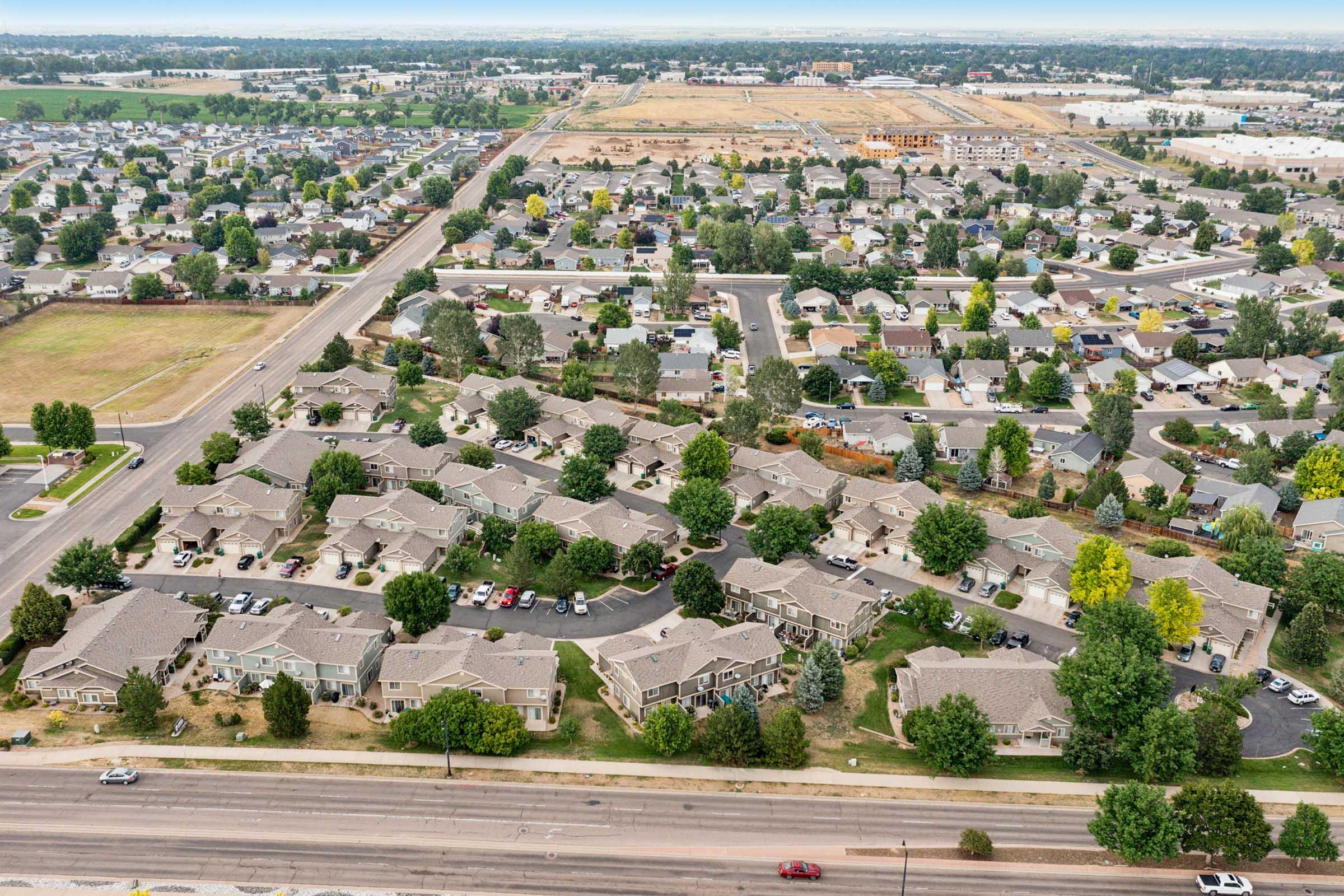 An aerial view of a suburban area featuring a variety of residential buildings, roads, and green spaces. The landscape includes clusters of homes with well-maintained lawns, along with a nearby empty lot and leveling for future development. The horizon shows a mix of urban and rural elements.