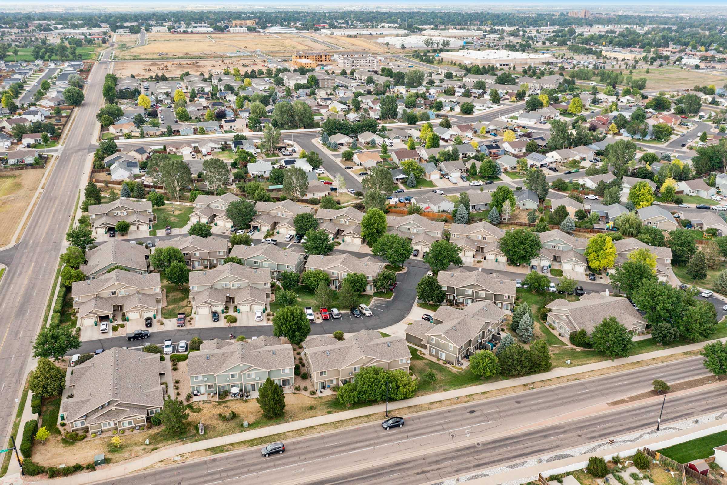 Aerial view of a suburban neighborhood featuring rows of houses with green lawns, trees, and parked cars. The surrounding area includes roads, empty lots, and a distant view of commercial buildings, suggesting a mix of residential and urban development.