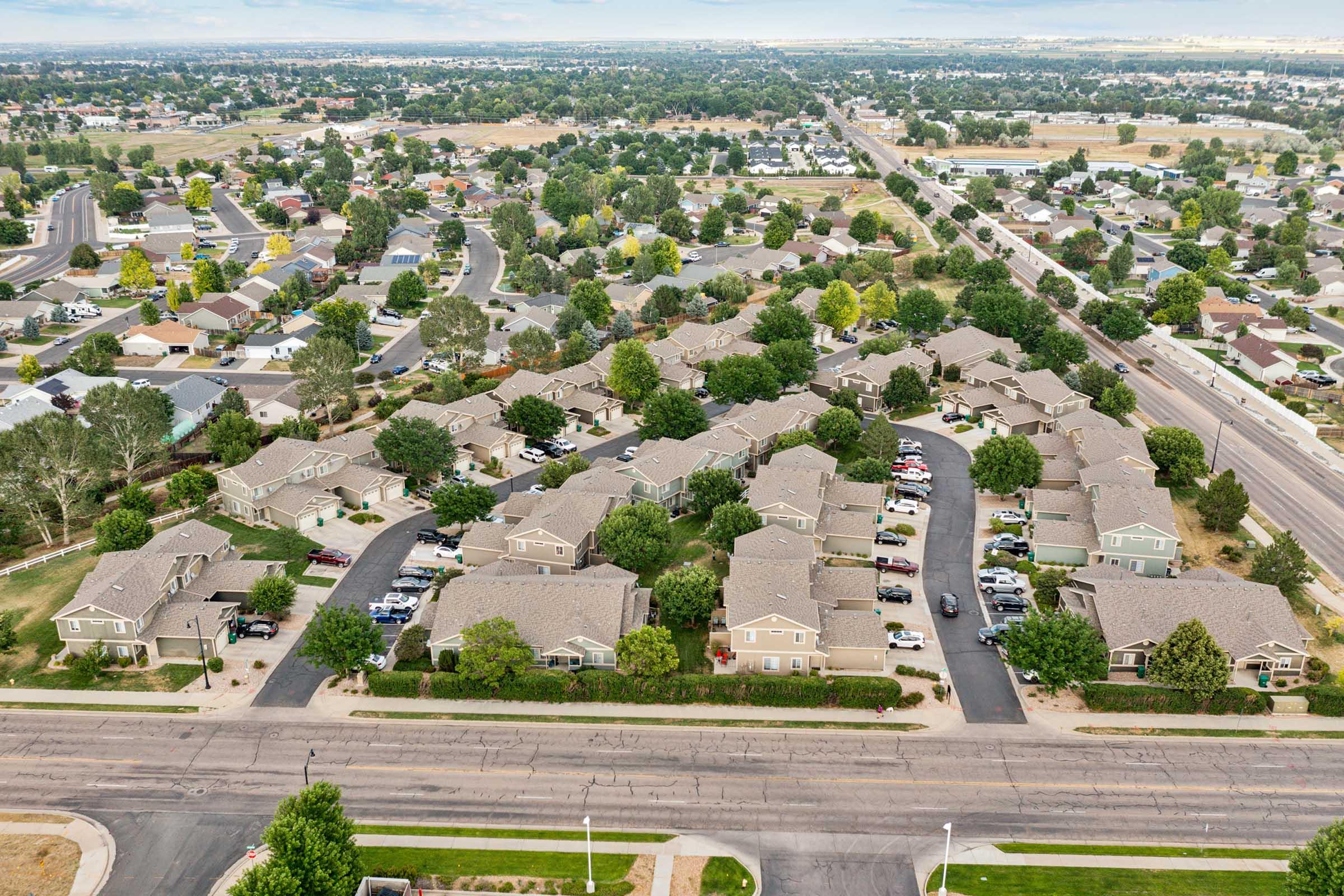 Aerial view of a suburban neighborhood featuring a layout of houses, with roads and parked cars. The area is surrounded by trees and greenery, showcasing a residential community with well-maintained lawns and pathways. In the background, additional homes and open land are visible under a partly cloudy sky.