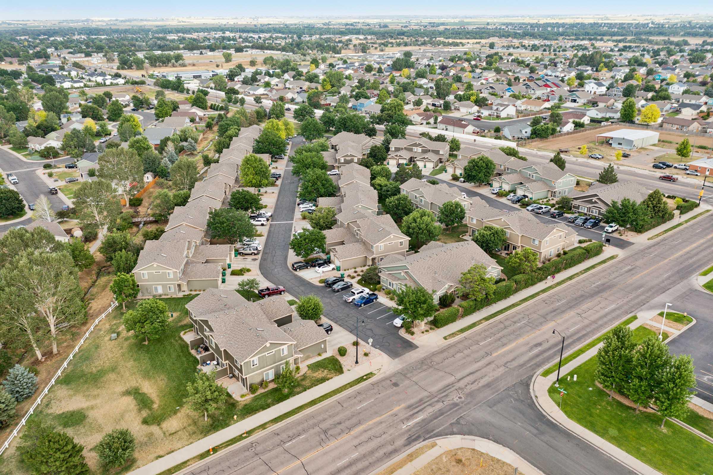 Aerial view of a suburban neighborhood featuring rows of houses with gray roofs, surrounded by green trees and open spaces. Several cars are parked in driveways and along the streets. The layout includes well-defined streets, grassy areas, and a distant view of more homes and open land.