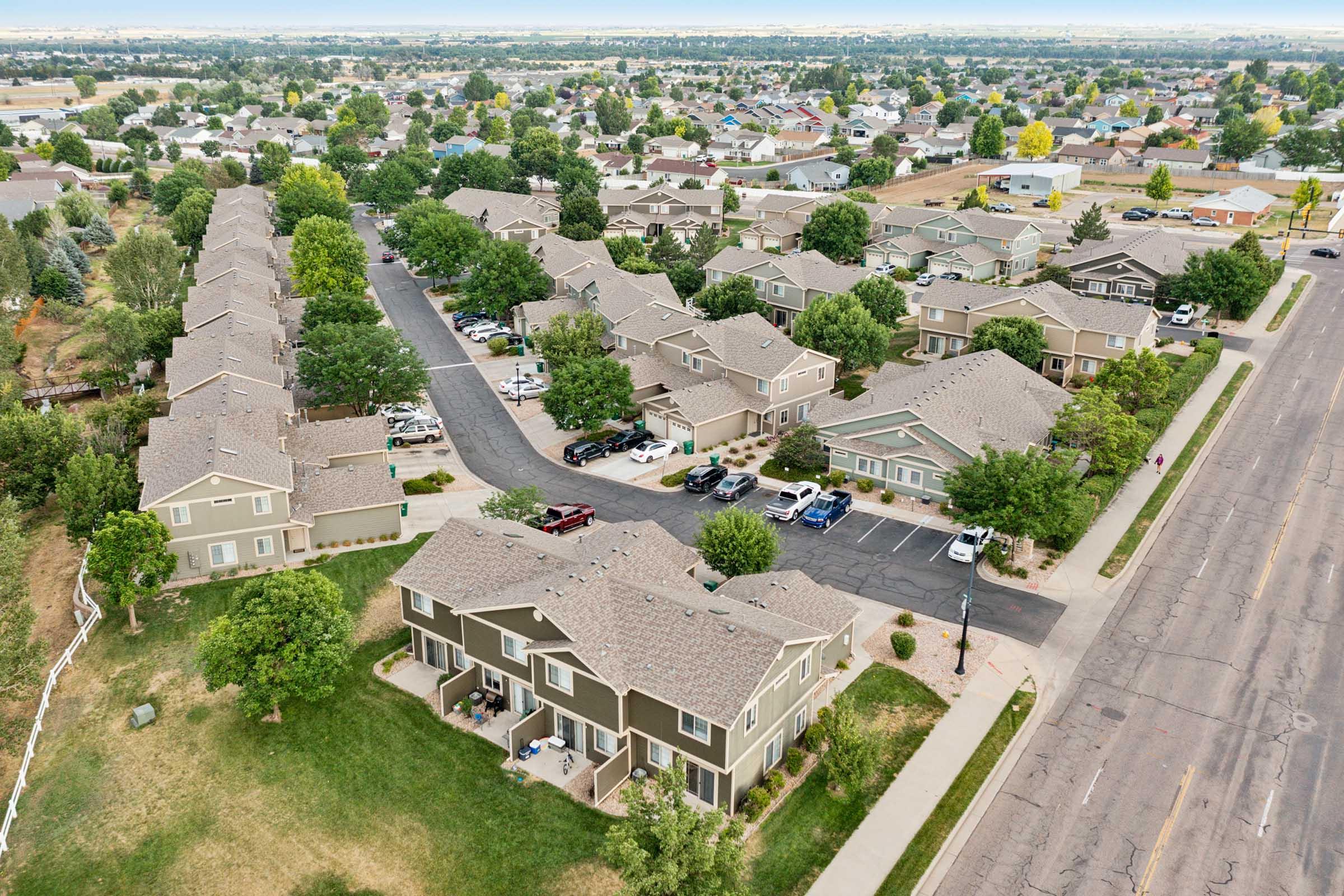 Aerial view of a suburban neighborhood featuring rows of houses, tree-lined streets, and parked cars. The landscape includes green lawns and open spaces, with additional homes visible in the distance.