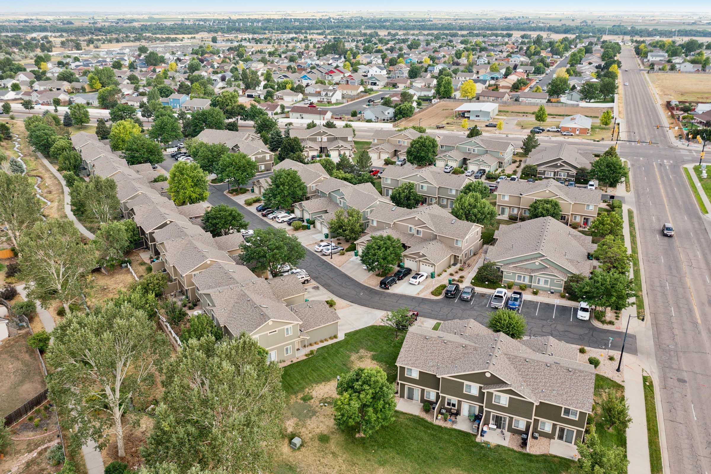 Aerial view of a suburban neighborhood featuring residential buildings with multiple houses arranged in a grid pattern, tree-lined streets, parked cars, and open green spaces. The background shows additional residential areas and farmland, under a clear sky.