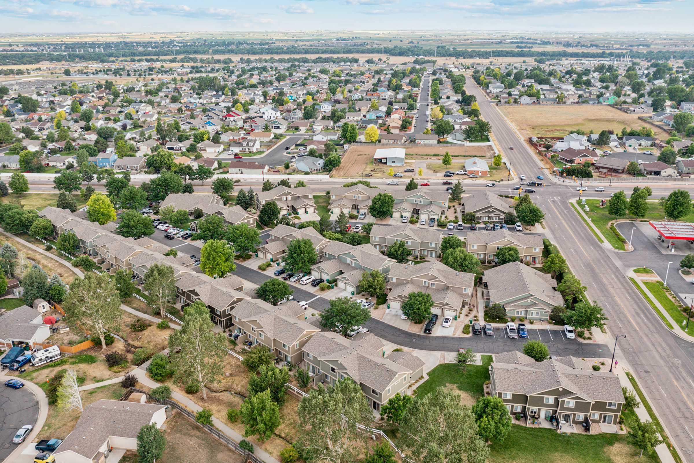 Aerial view of a suburban neighborhood with clusters of houses, green trees, and streets. The image shows well-maintained residences, parking areas, and a vast landscape in the background, highlighting a mix of developed and open land.