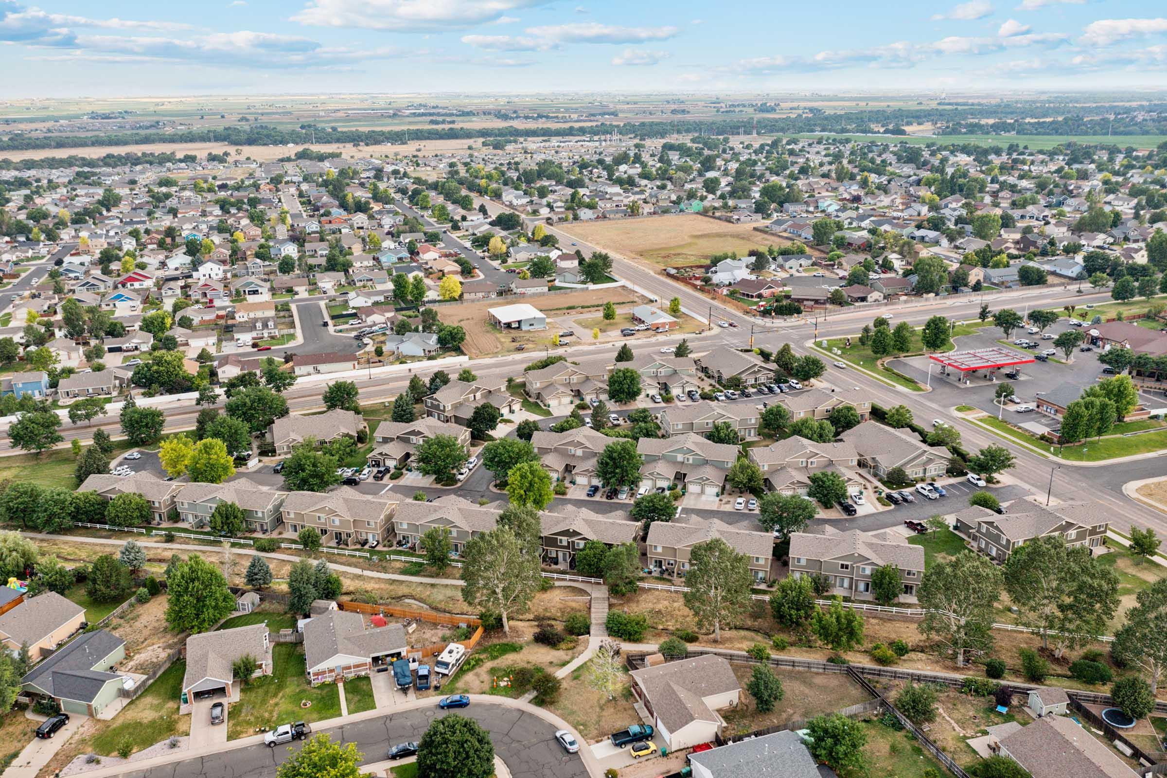 Aerial view of a suburban neighborhood featuring rows of residential homes with varying roof styles, ample trees, and green spaces. In the background, fields and a few commercial buildings are visible, along with a road lined with parked cars. The scene showcases a mix of urban and rural landscapes under a clear blue sky.