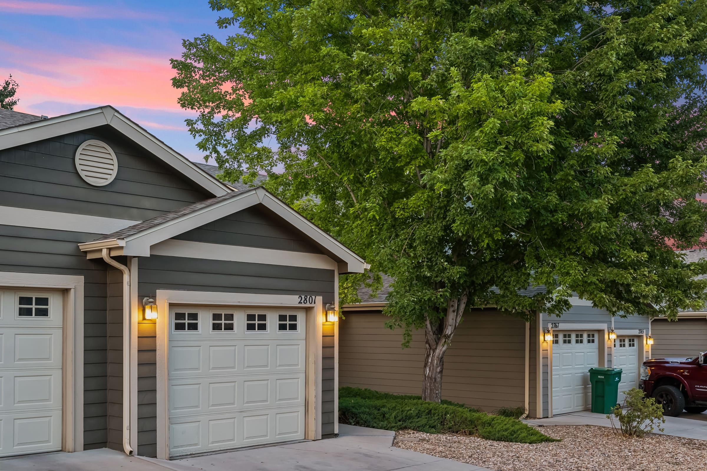 A suburban neighborhood scene featuring two attached garages with white doors, a tree nearby, and colorful sunset skies in the background. Decorative landscaping with rocks is visible in the foreground, alongside green trash bins by the driveway.