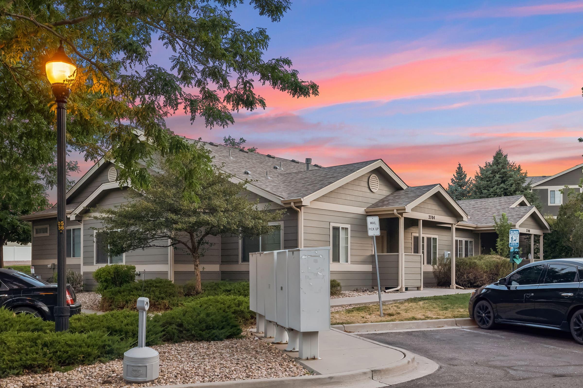 A residential building with light brown siding, surrounded by green shrubs and trees, located in a quiet neighborhood. A streetlamp illuminates the area at dusk, with a colorful sunset sky in the background. Several mailboxes are visible near the entrance, and parked cars are seen nearby.