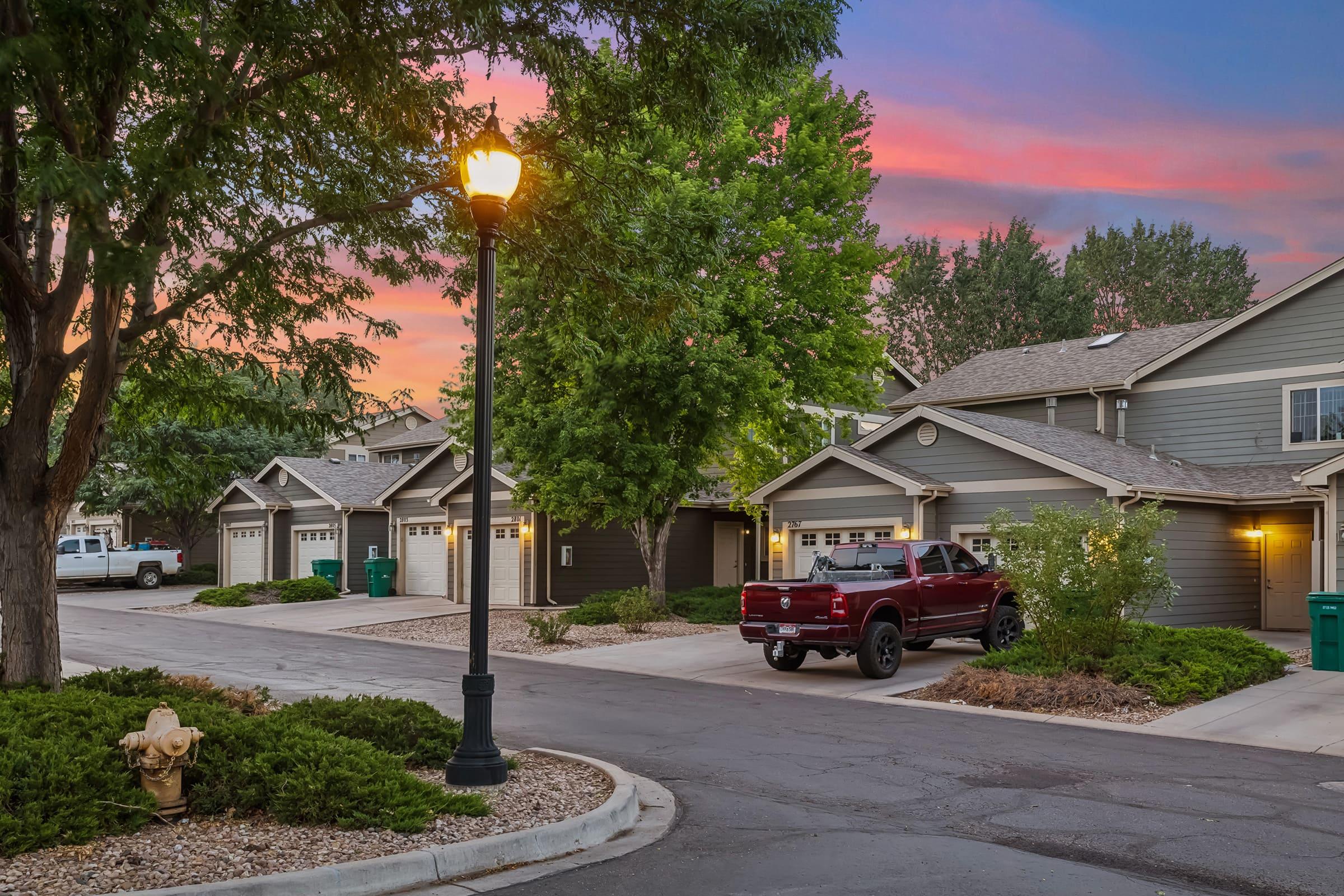 A peaceful suburban street at sunset, featuring several houses with garages. A red pickup truck is parked in front of one house, while a lamppost illuminates the scene. Lush green trees and shrubs line the street, creating a serene atmosphere against a colorful sky filled with pink and blue hues.