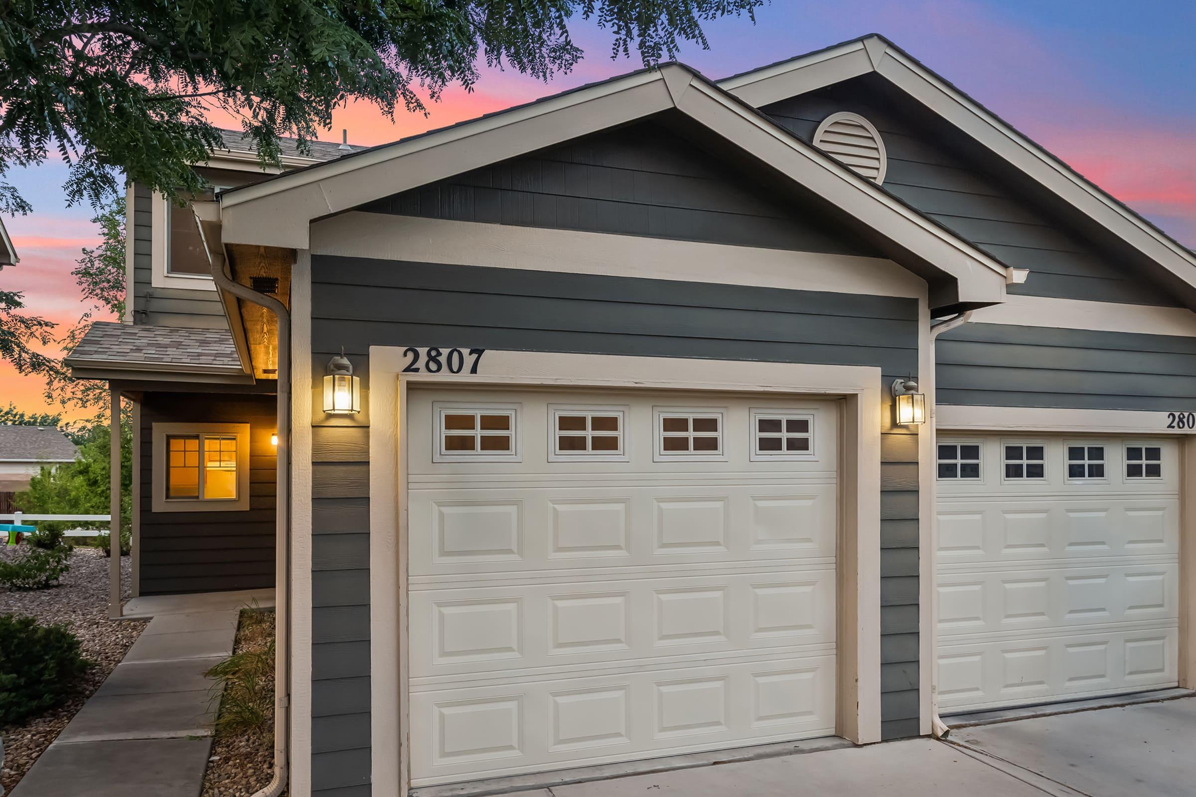 A modern two-story home with a dark gray exterior and white trim, featuring a double garage door. The driveway is lined with stones, and there are decorative lights beside the garage. The sky in the background is painted with shades of pink and purple, indicating sunset.