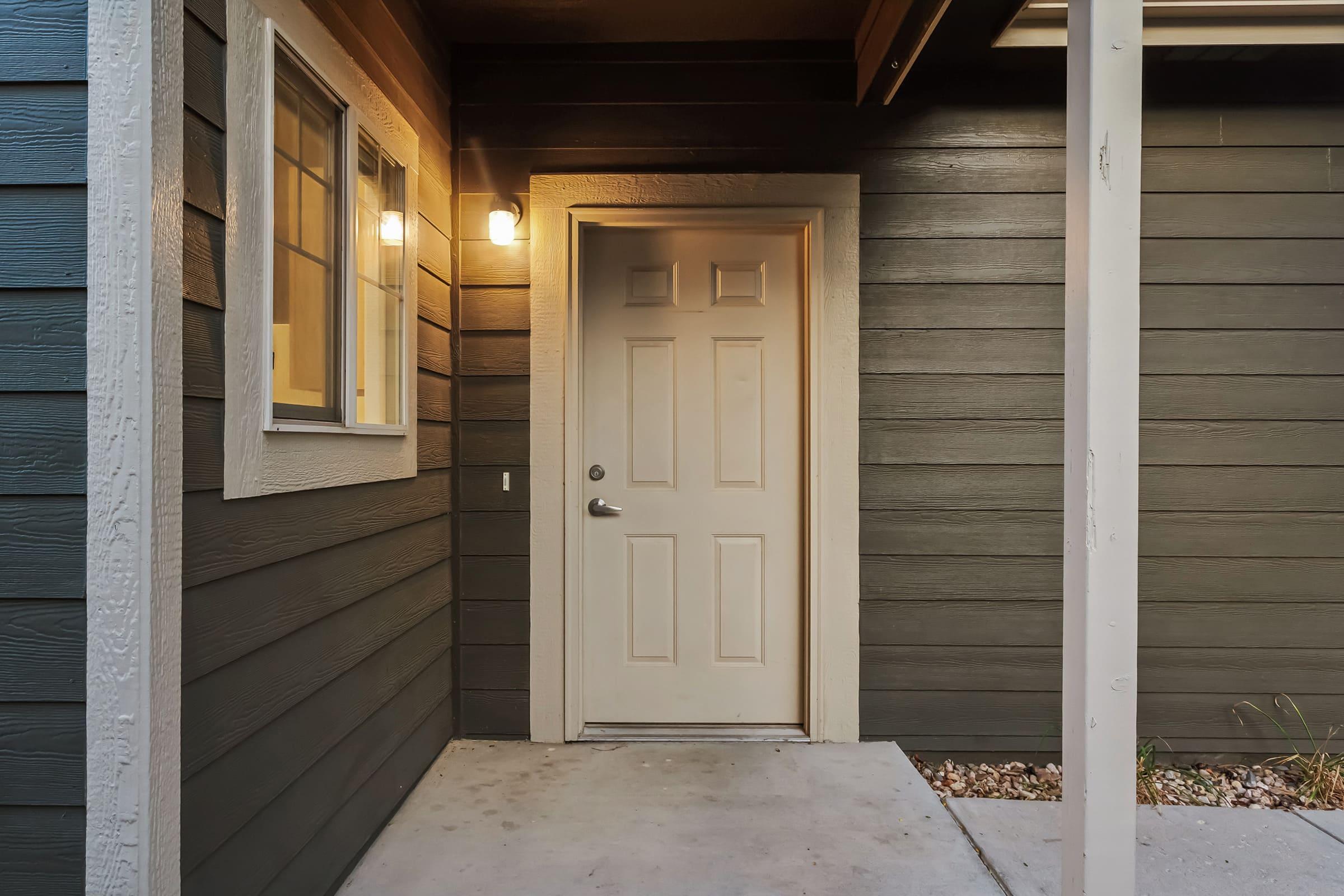 Exterior view of a house entrance featuring a light-colored door, adjacent window, and dark wooden siding. The area is well-lit by a wall-mounted light fixture, with a small porch and concrete flooring. Decorative stones are placed near the entrance.