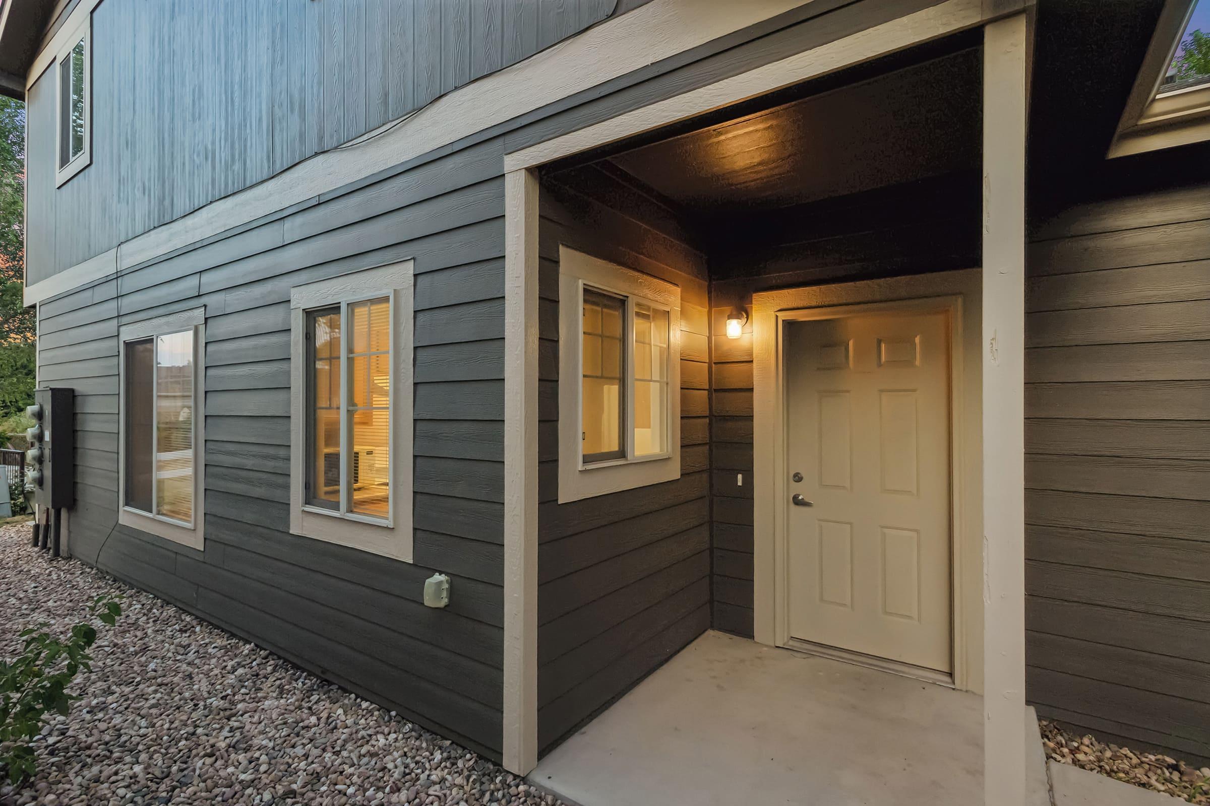 Exterior view of a modern house entrance featuring a light-colored door and windows with warm lighting. The walls are dark wood paneling, and the area is surrounded by small stones. A light illuminates the entrance, creating a welcoming atmosphere.