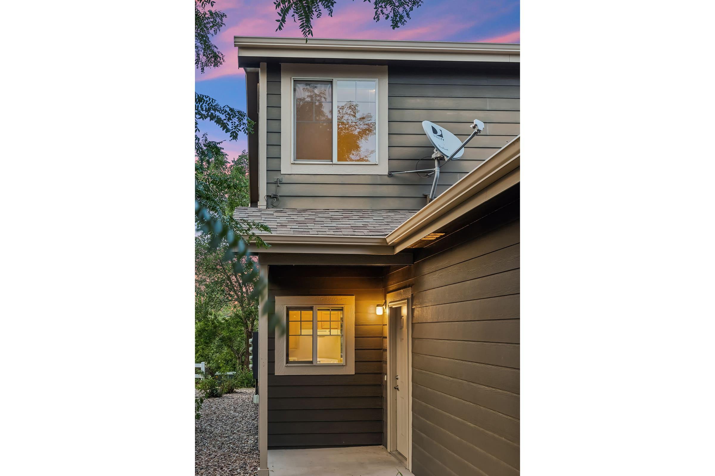A modern two-story house with dark wood siding. The house features a front door with a warm light above it, and a satellite dish mounted on the roof. A window on the upper level reflects the sunset, creating a warm ambiance. Surrounding greenery and pebbled ground complete the scene.