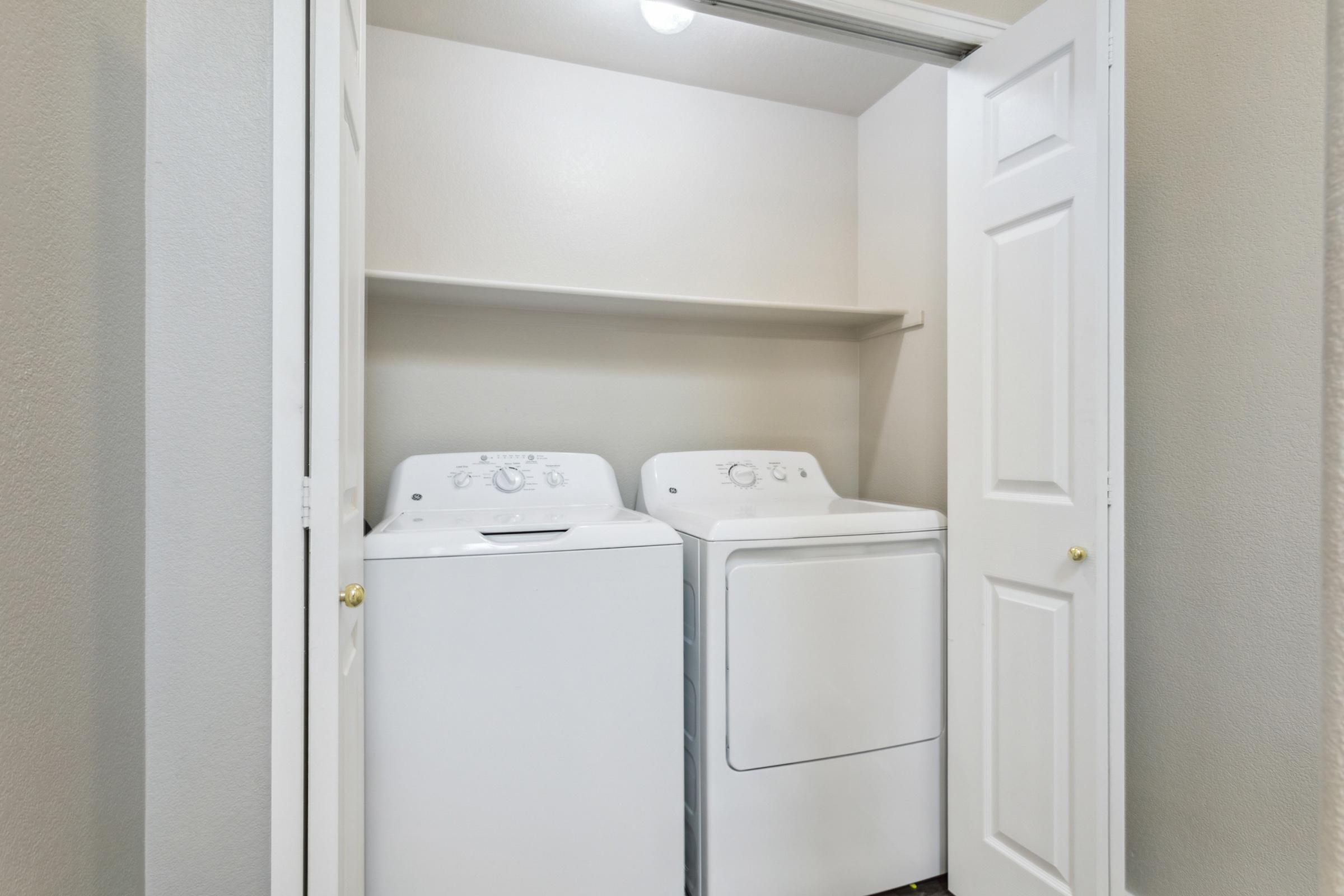 A clean laundry closet featuring a stacked washing machine and dryer side by side. The appliances are white and located under a shelf, with slightly open double doors revealing a neutral-colored wall. The lighting is bright, indicating a well-lit space.