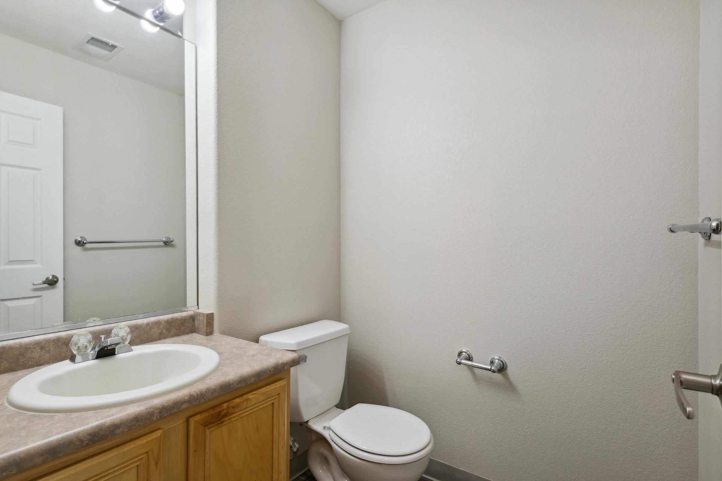 A small bathroom featuring a beige wall, a single sink with a wooden cabinet, a mirror above the sink, a white toilet, and a towel bar. The space is well-lit, with a door and a light fixture visible.